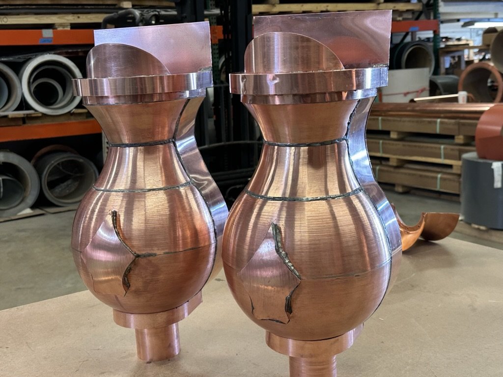 Two rounded copper leader heads, set on a workshop table with industrial shelving and pipes in the background.