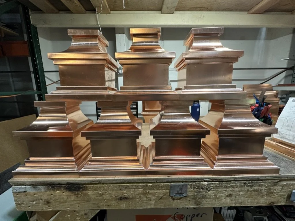 Stacked copper leader heads / scupper boxes arranged on a workbench in a workshop.