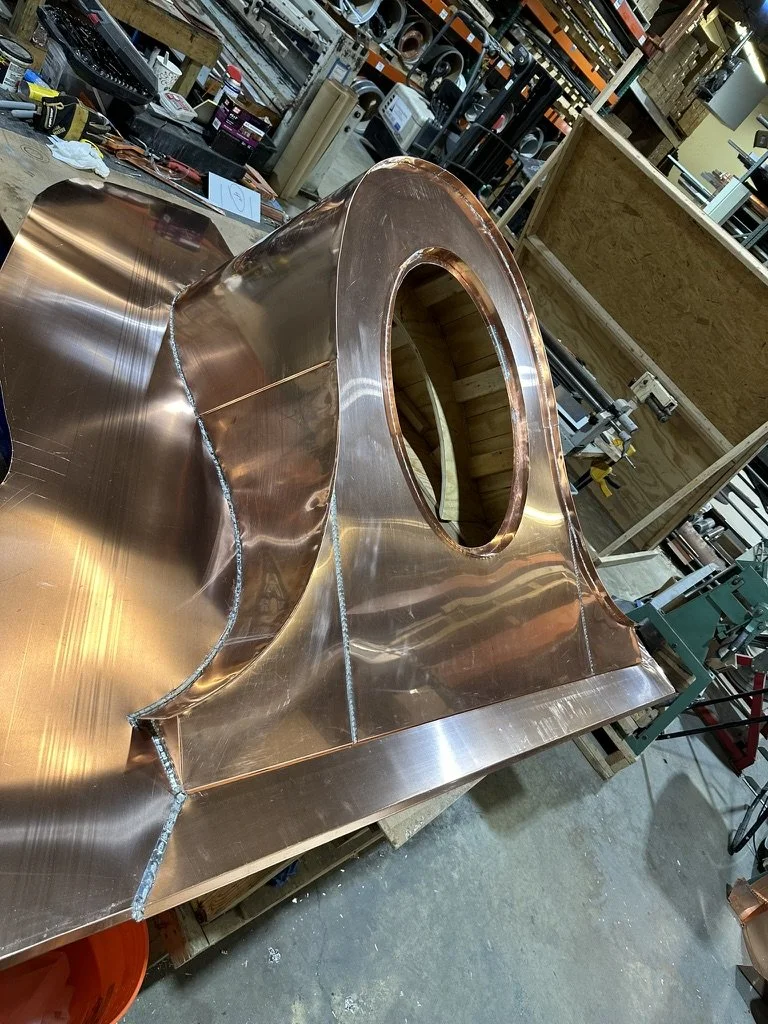 A large, shiny copper dormer in progress on a worktable in a workshop, with tools and shelves in the background.