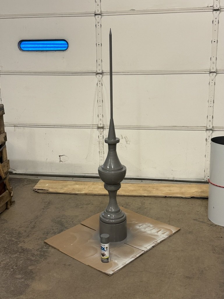 A tall, decorative metal finial painted gray, standing upright on a piece of cardboard in a garage