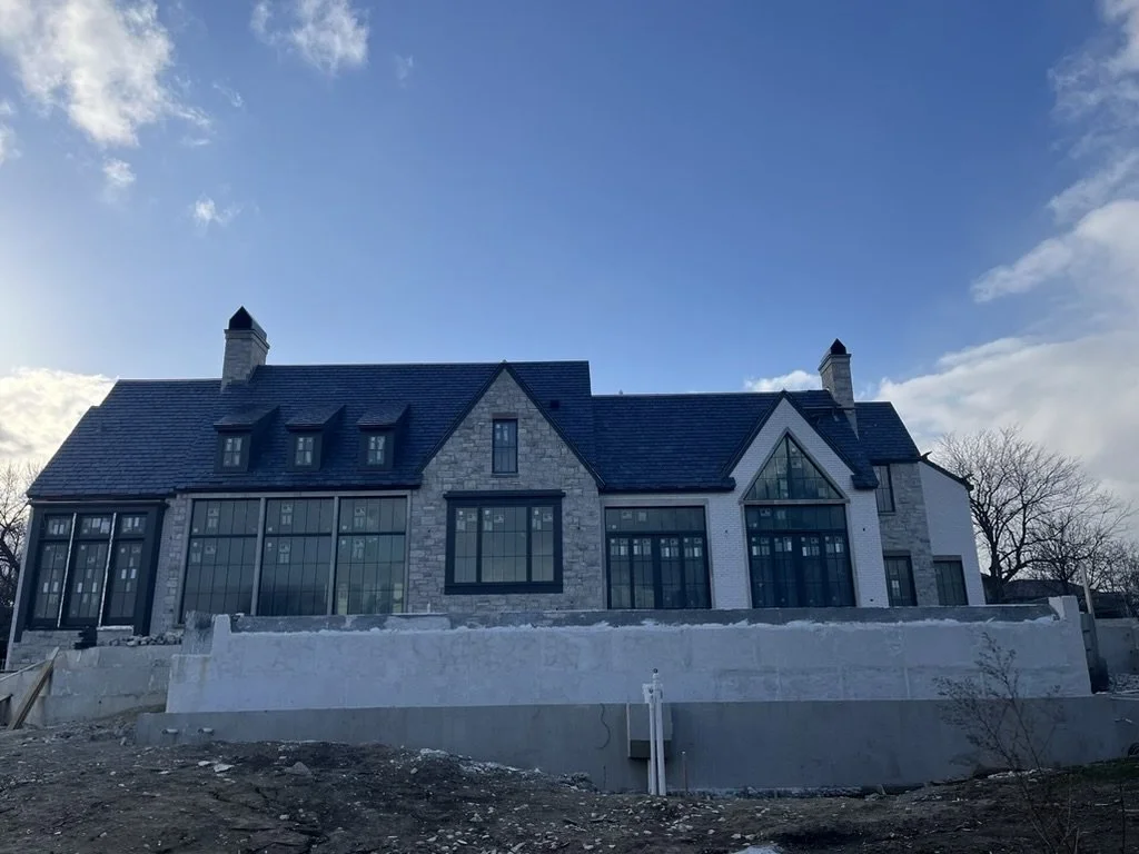 A large house with a stone and white exterior, multiple large windows, and a steep black roof, under a partly cloudy sky.