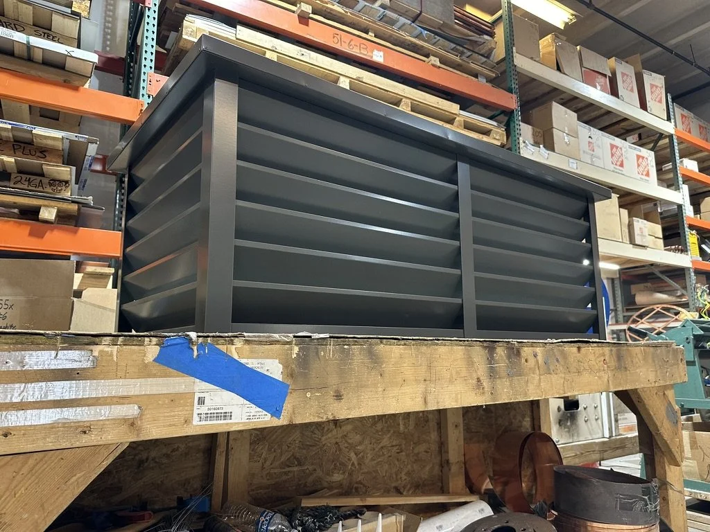 A black metal louvered chimney with horizontal slats placed on a wooden workbench in a warehouse, with shelves full of boxes and supplies in the background.