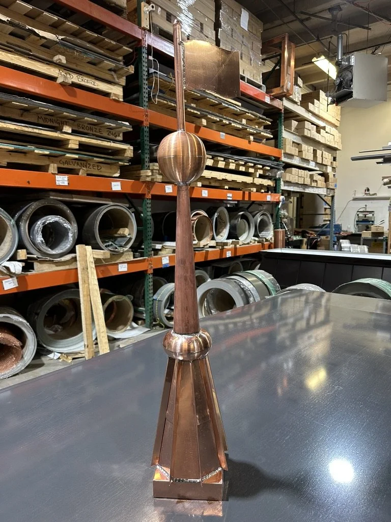 A tall, copper finial / spire sculpture resembling a weather vane or abstract weathervane with a spherical top, a long vertical rod, and a pyramid-shaped base, placed on a metallic work table in a workshop or warehouse with shelves of metal parts.