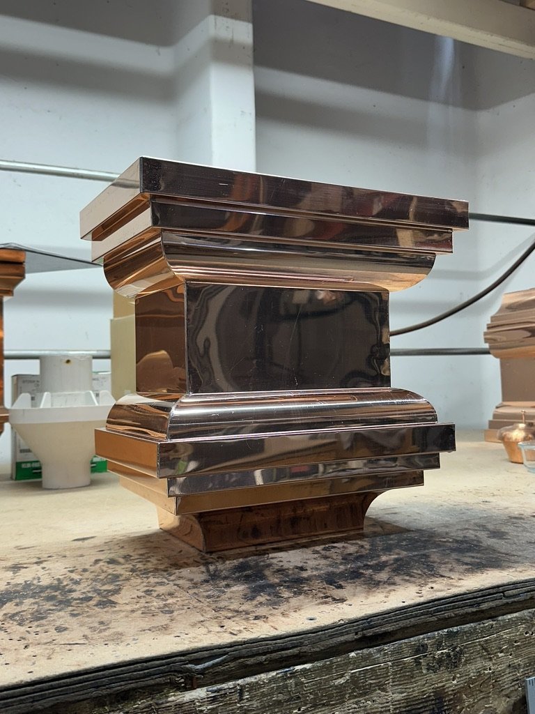 A shiny metallic, ornate square-shaped leader head / scupper box with curved edges, placed on a workbench in a workshop.