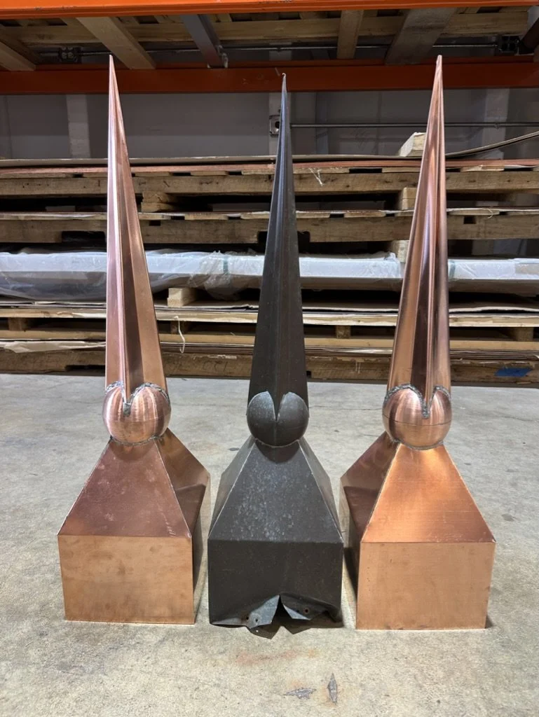 Three metal sculptures resembling tall, pointed spires with rounded bases, two are new copper and one is dark gray, arranged symmetrically on a warehouse floor. One is old and two are historical reproductions.