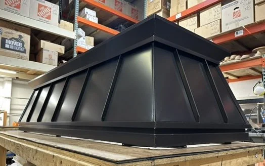 Large black metal chimney shroud with a slanted design placed on a wooden pallet in a warehouse.