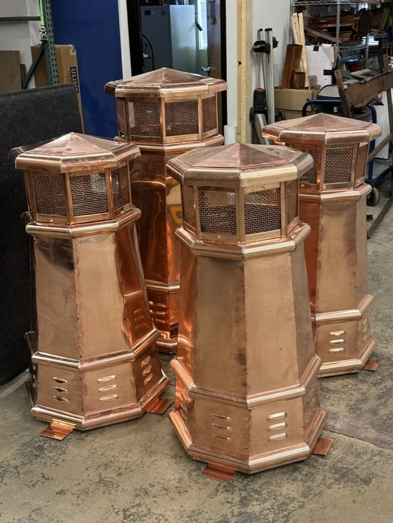 Four large, polished copper chimney shrouds / pots with octagonal shapes, mesh windows at the top, and louvered vents near the base, placed on a workshop floor.