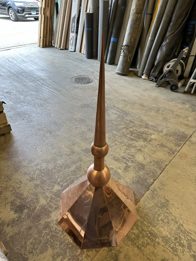A large, shiny copper-colored spear-shaped finial / spire sculpture with a broad, multi-faceted base, situated inside a workshop.