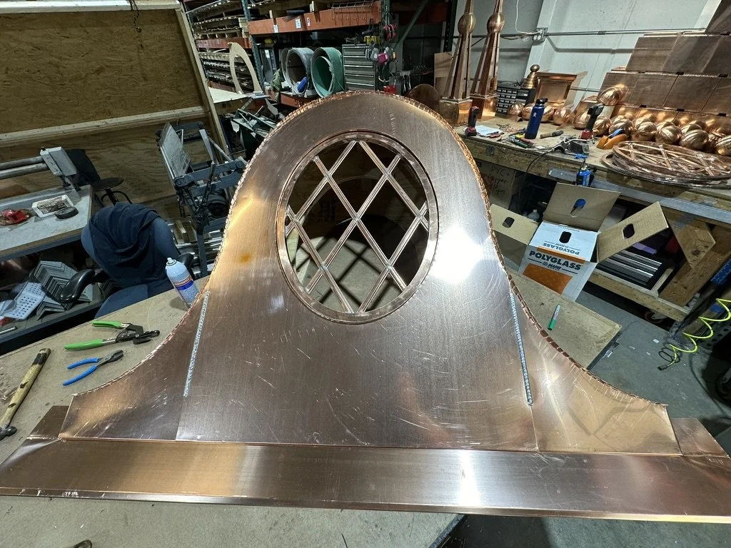 A copper dormer with a large circular opening and grid patterned window inside, placed on a workbench in a workshop surrounded by tools and other metal objects.