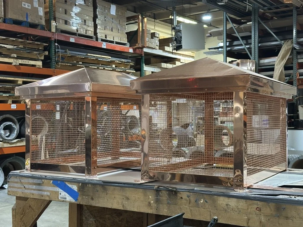 Two copper chimney shrouds with mesh sides and sloped roofs are on a workbench in a warehouse.