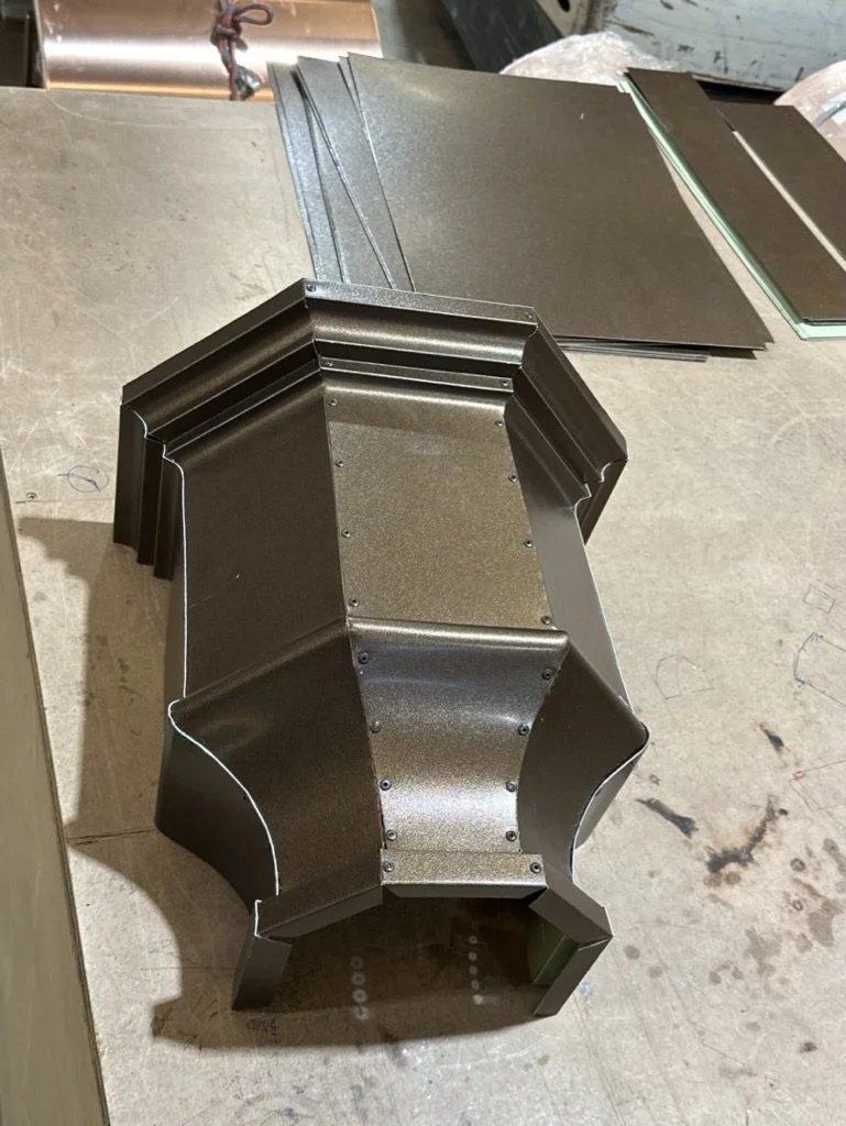 A black metal leader head / scupper box, shaped like a small container or casing, placed on a workbench with sheets of black material and a copper pipe in the background.