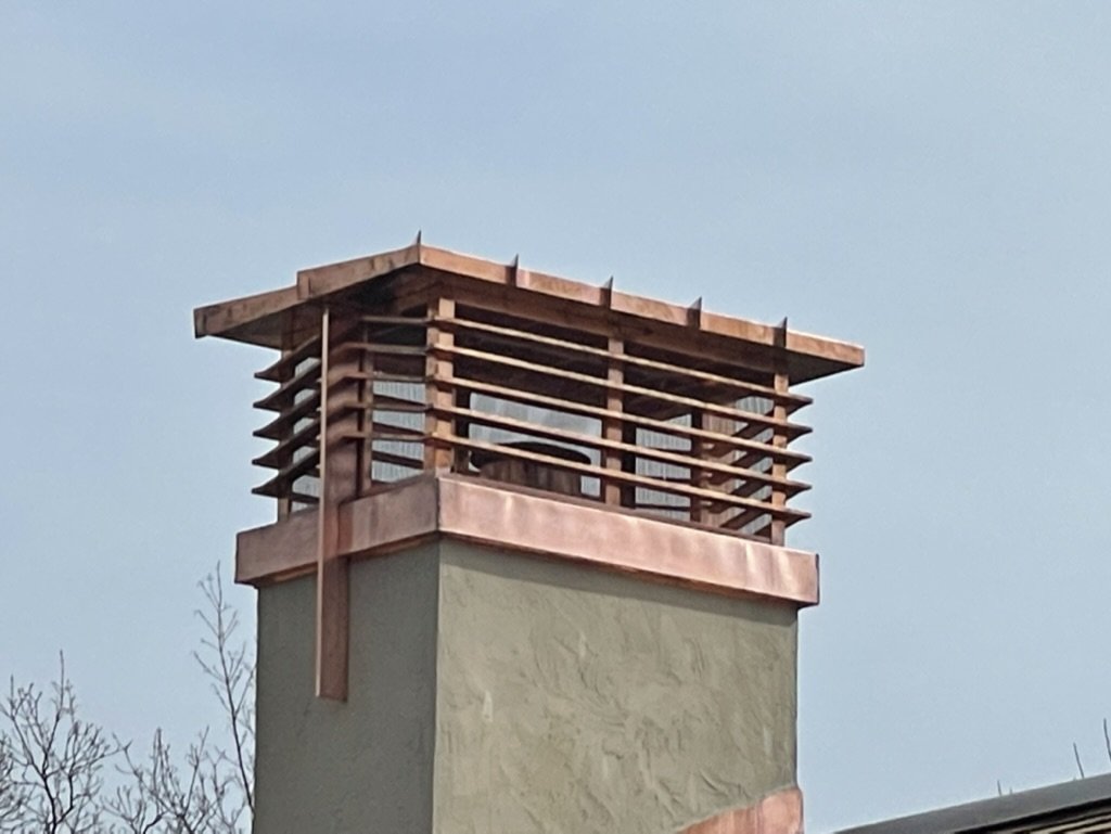 Close-up of a chimney with a midcentury-inspired copper shroud and weather-resistant louvred vents on top of a building, set against a clear sky.