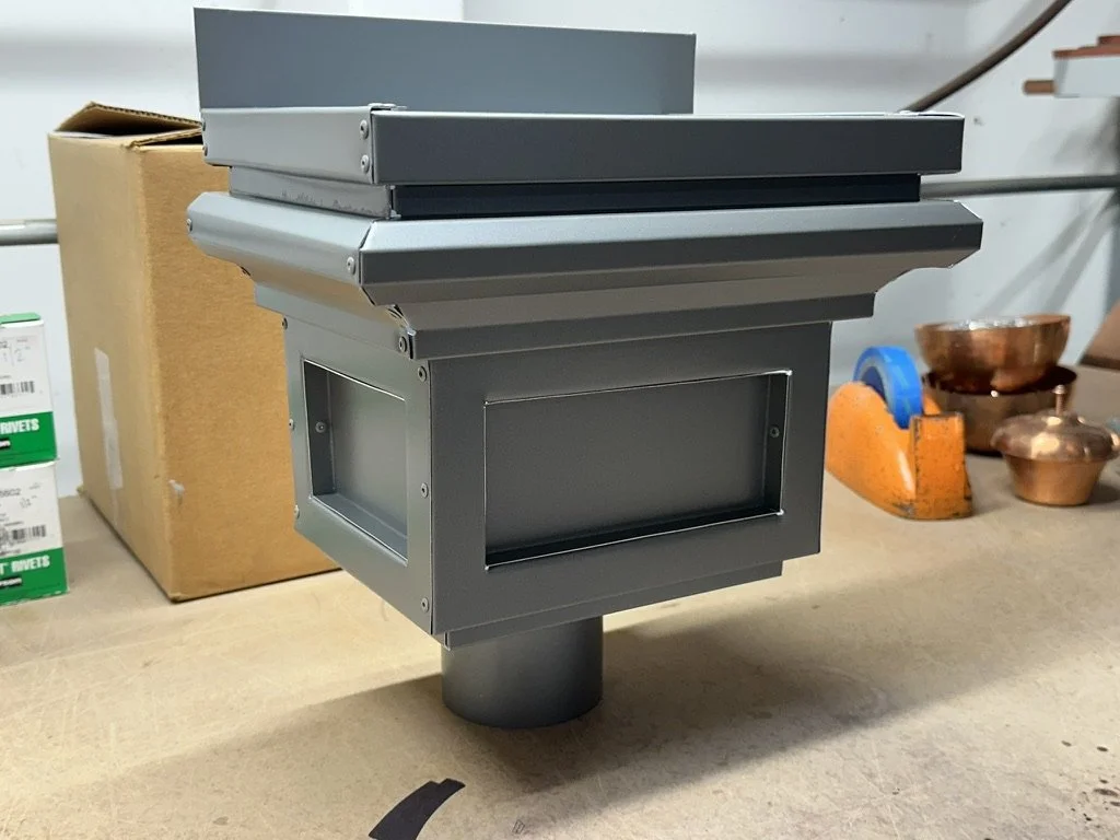 Gray aluminum leader head / scupper box with a rectangular opening, mounted on a stand, with a cardboard box and copper bowls in the background.