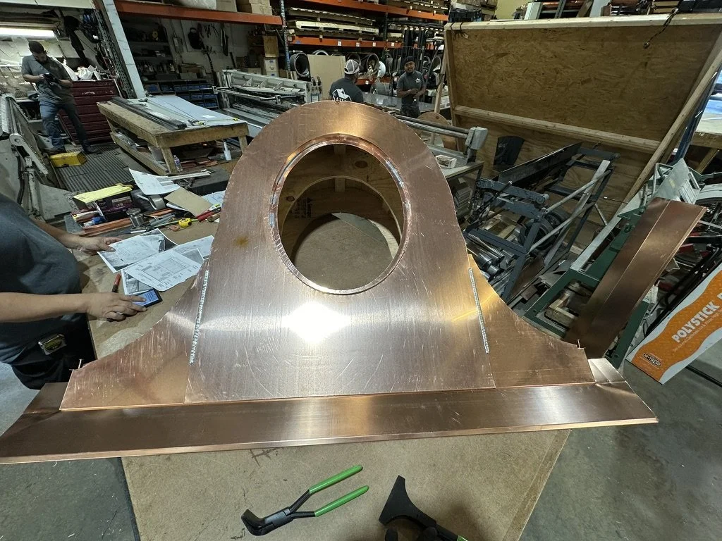 A large, copper dormer component with a circular hole in the center, placed on a worktable in a manufacturing workshop. The workshop has various tools, equipment, and workers in the background.