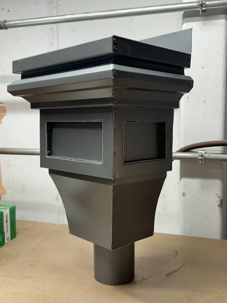An aluminum leader head / scupper box with a tapered lower section, sitting on a wooden surface in a workshop or factory setting.