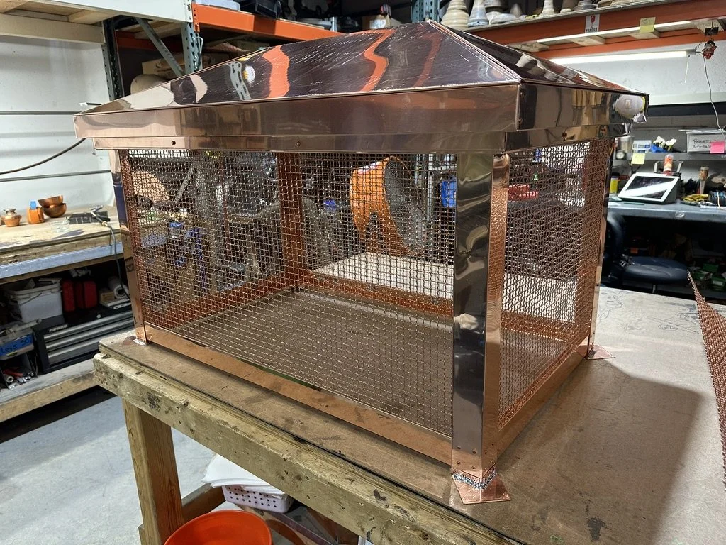 A copper chimney shroud with a pyramid-shaped roof and mesh sides, placed on a workbench in a workshop.