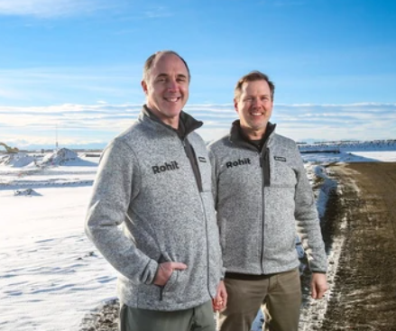 Two men standing on a snowy landscape wearing Rohit branded gray jackets, smiling at the camera, with a clear blue sky in the background.