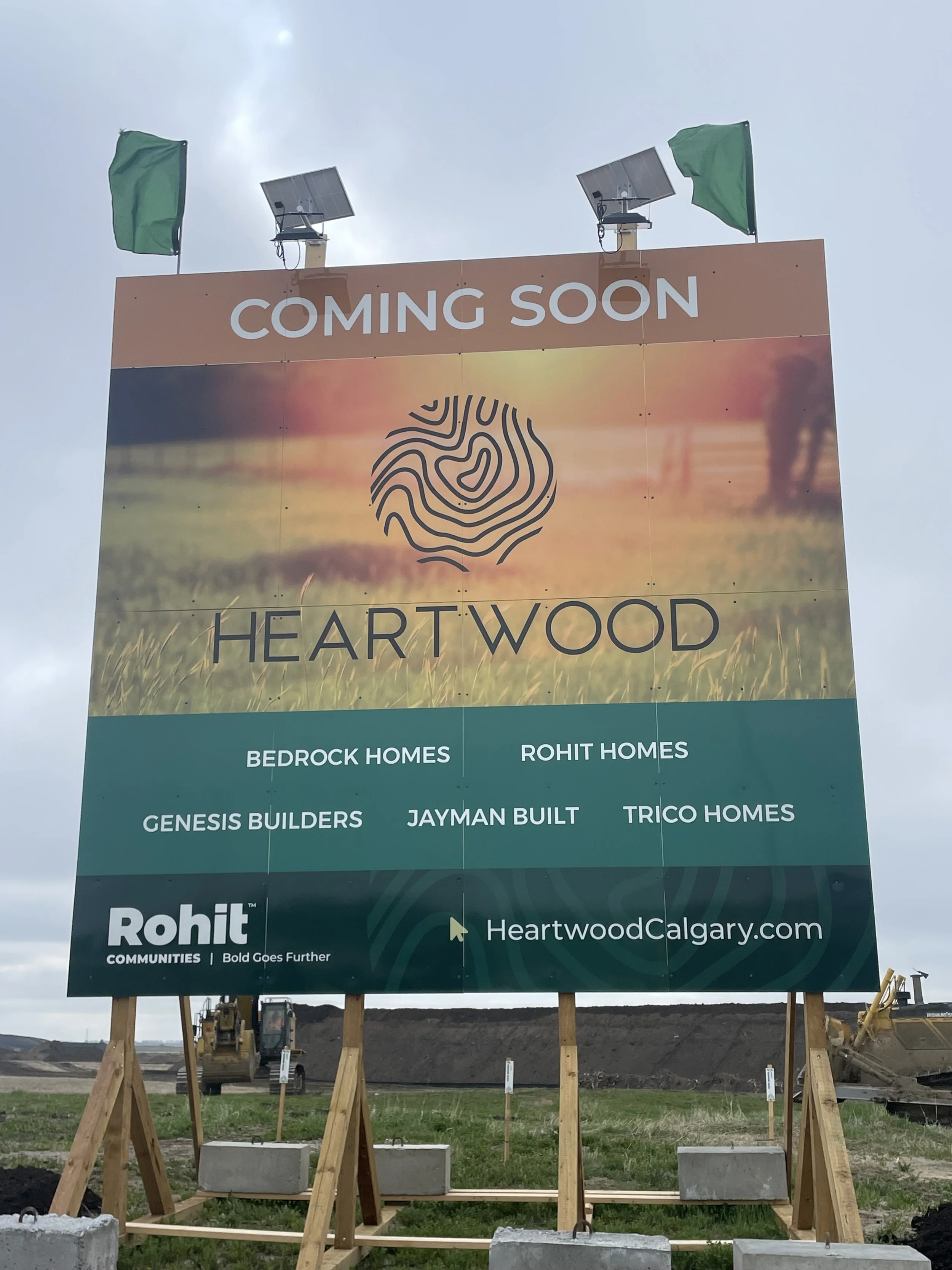 Billboard advertising 'Heartwood,' a new development, with information about associated building companies including Bedrock Homes, Rohit Homes, Genesis Builders, Jayman Built, and Trico Homes. Website link: HeartwoodCalgary.com. Background shows construction site with earth-moving equipment.