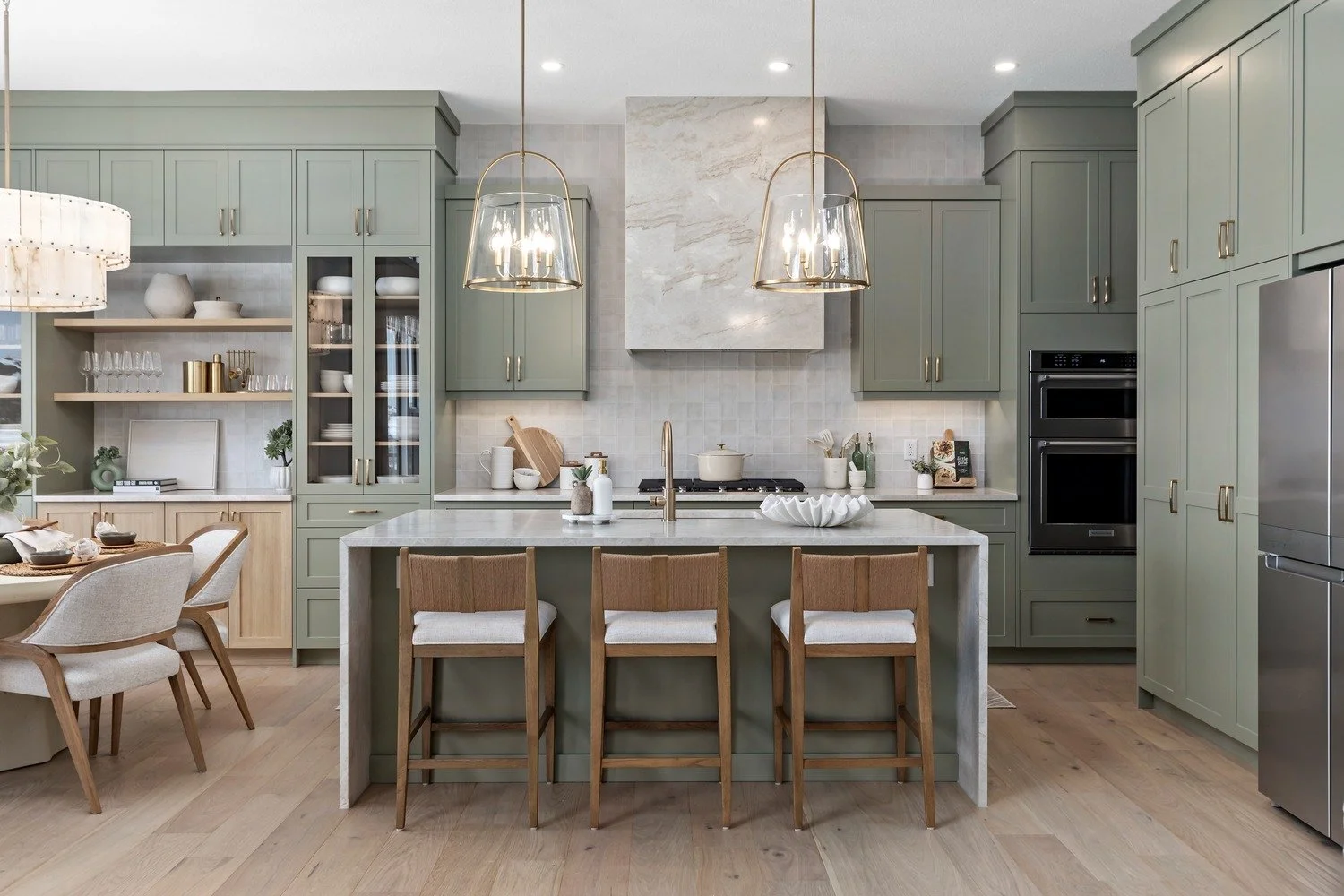 Modern kitchen with green cabinetry, marble island, pendant lights, and beige chairs.