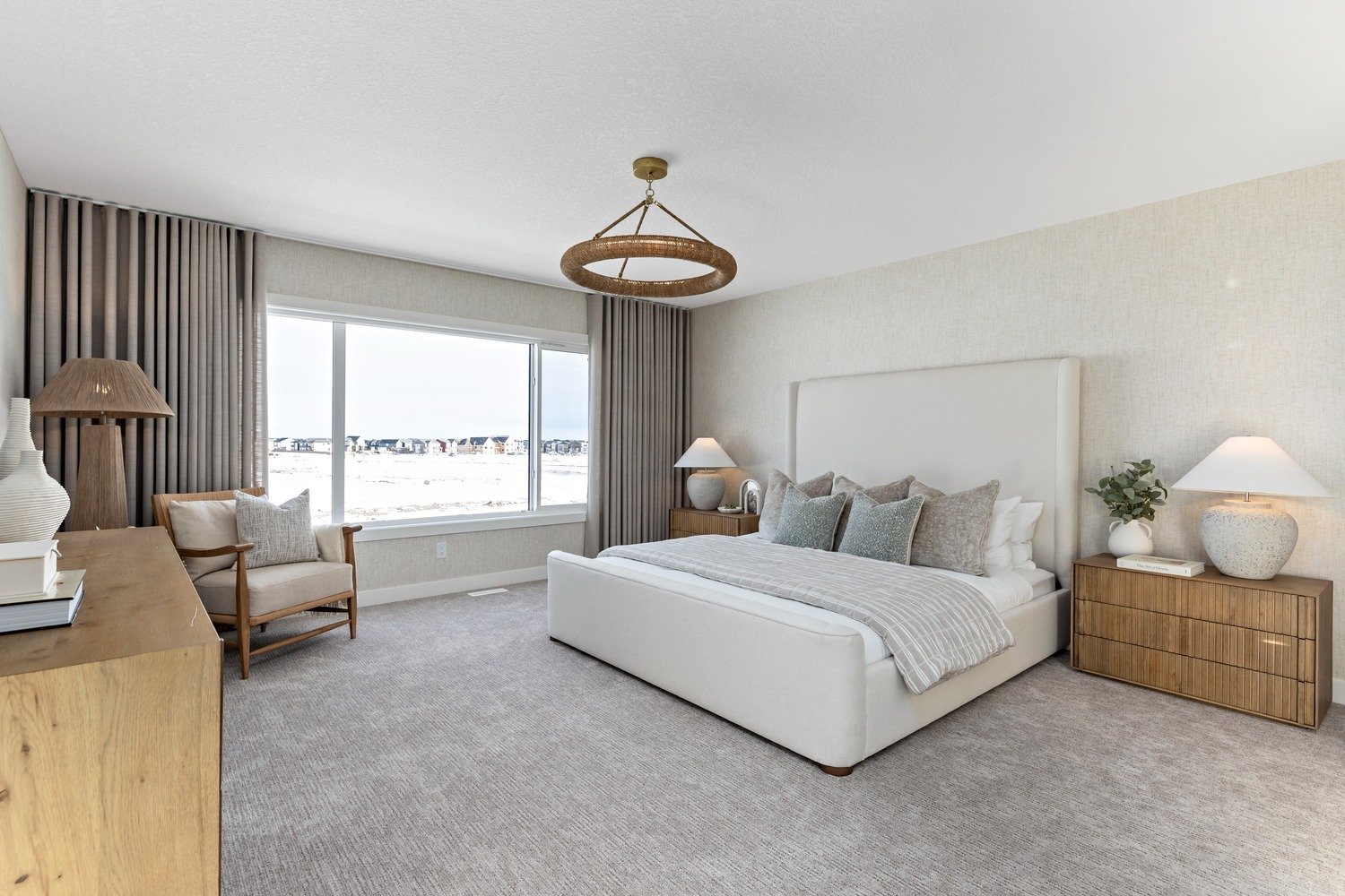 A bedroom with a large window showing snow outside, a white bed with multiple gray pillows, wooden nightstands with lamps, a beige armchair, and a wooden dresser with decorative vases and books.