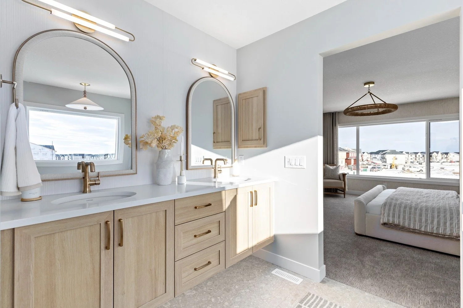 Modern bathroom with dual sinks, large mirrors, and natural light, overlooking a bedroom with large windows and a bed.