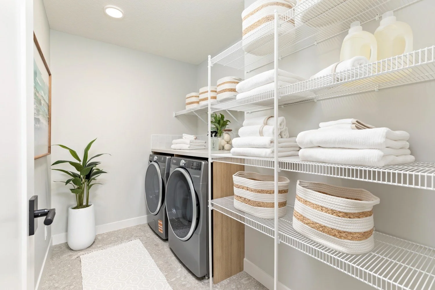 A laundry room with a washing machine and dryer, white wire shelves filled with neatly folded white towels and woven baskets, and a potted green plant in a white vase.