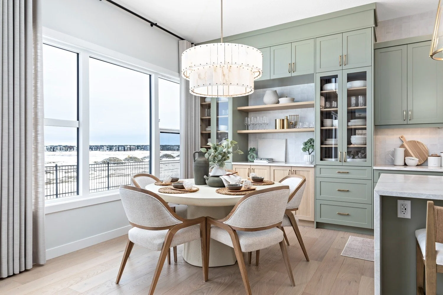A dining area with a round table and six upholstered chairs in a modern kitchen with large windows, light green cabinetry, open shelves, and a chandelier overhead.