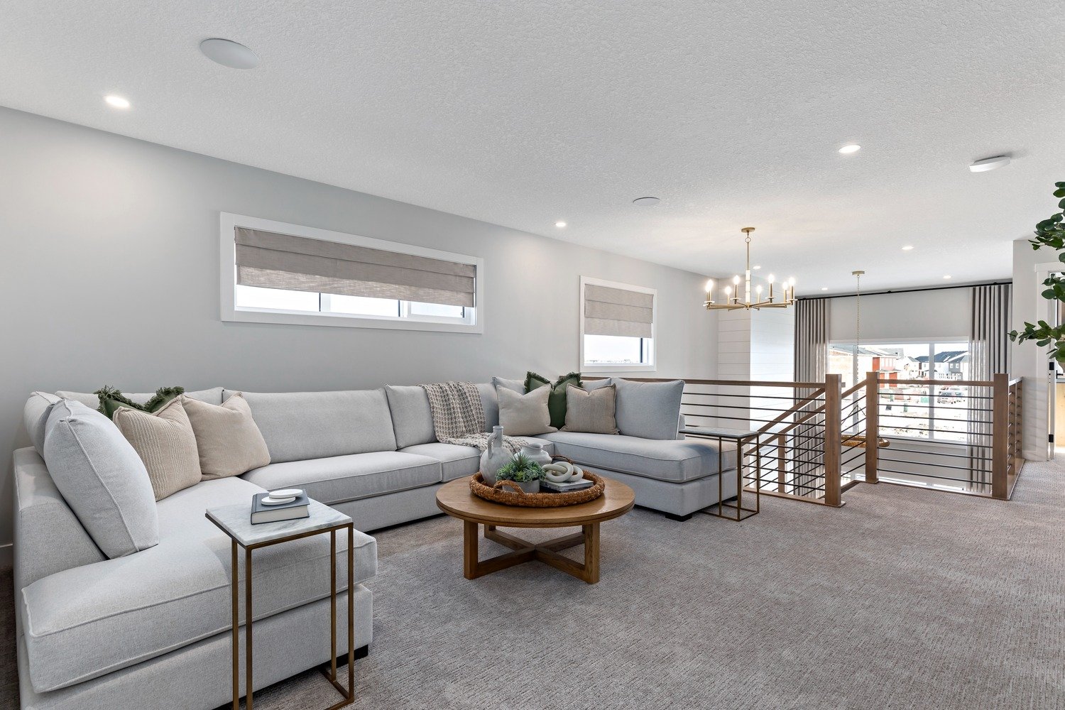 Living room with light gray sectional sofa, beige pillows, wooden coffee table, side table with books, window blinds, modern chandelier, staircase with wooden handrail, windows showing an outdoor view