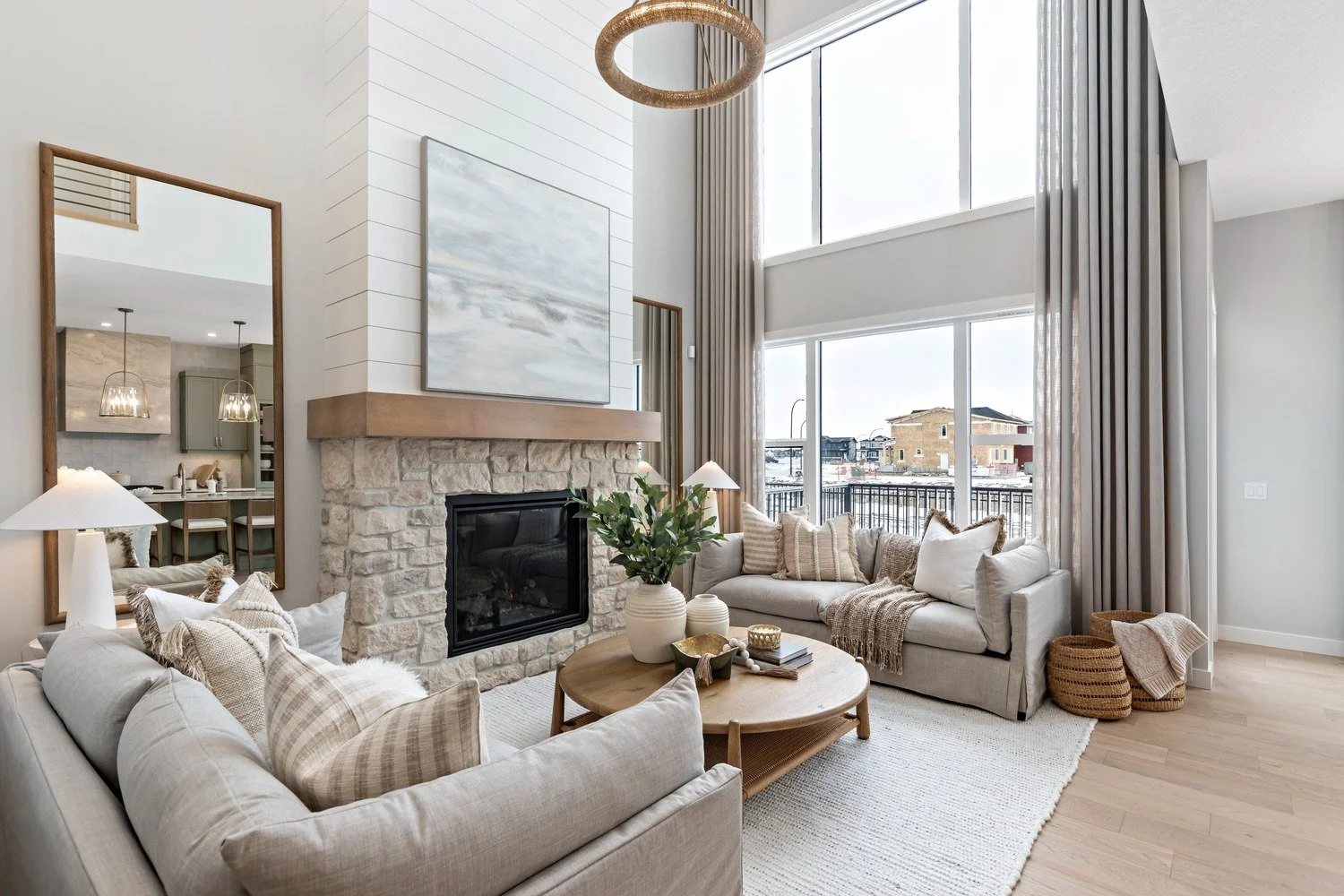 Modern living room with a two-story ceiling, large windows, a stone fireplace, and neutral-toned furniture and decor.