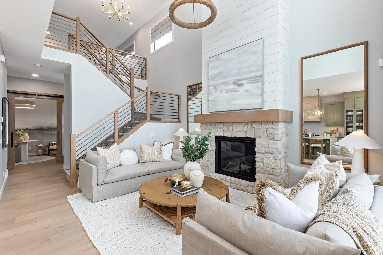 Living room with a beige sofa, decorative pillows, a wooden coffee table with decor and books, a fireplace with a stone surround and a large abstract painting above, a mirror reflecting part of the room, a staircase with wood and metal railing, and a
