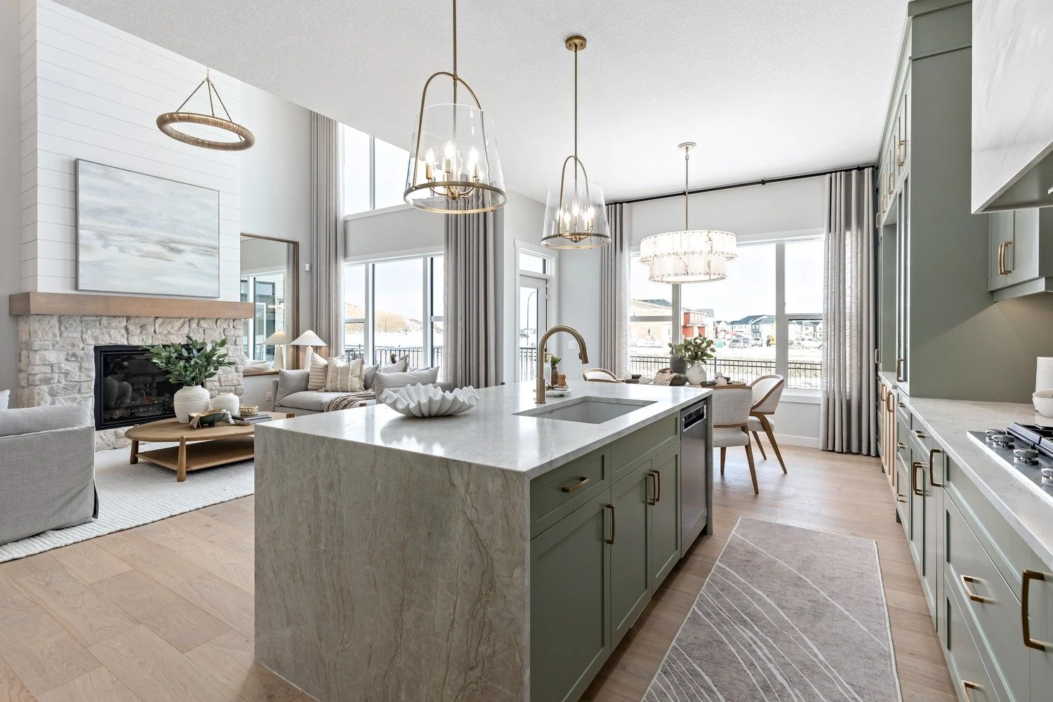 Modern open-concept living room and kitchen with a large island, light green cabinets, and a dining area near large windows with curtains, in a bright home with high ceilings and contemporary decor.