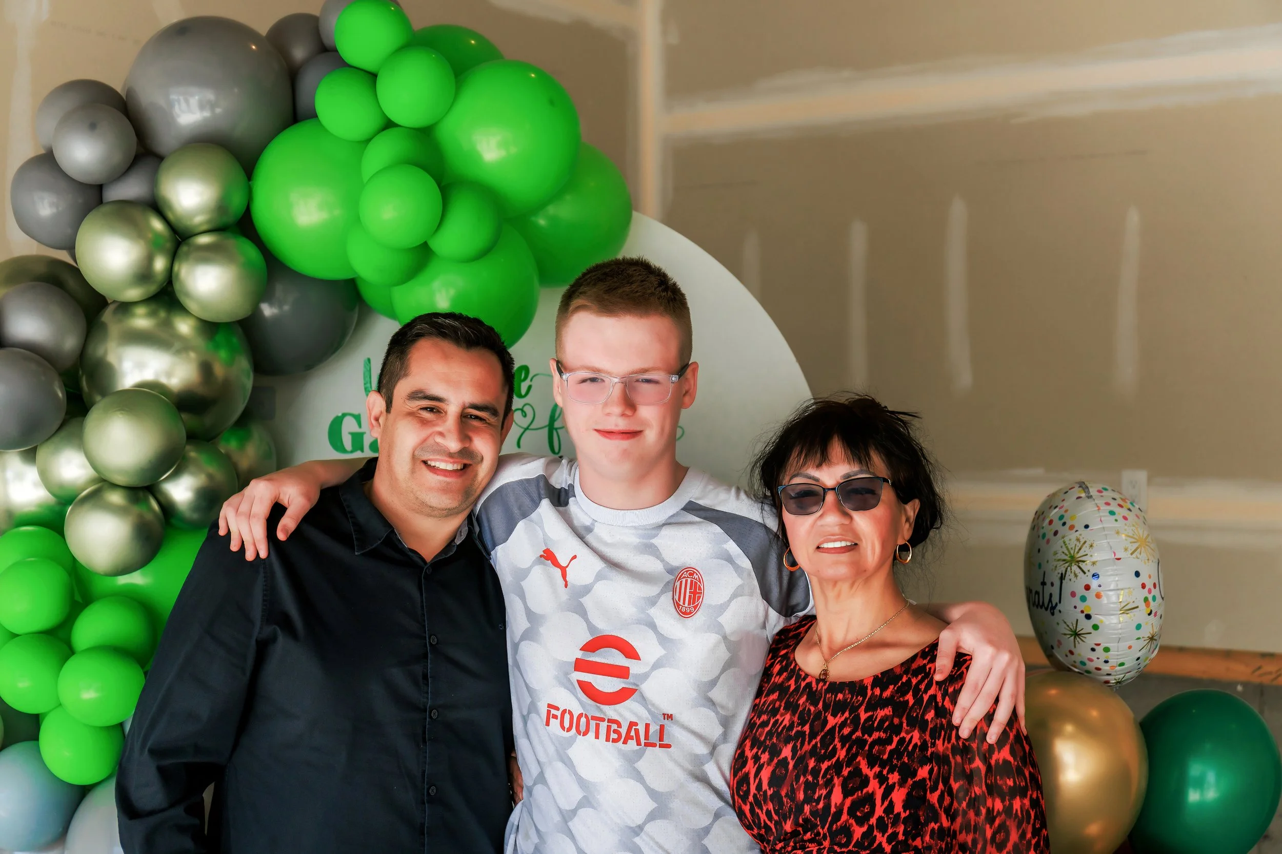 Three people smiling, standing in front of a green and gray balloon arch, with one person wearing an AC Milan soccer jersey.