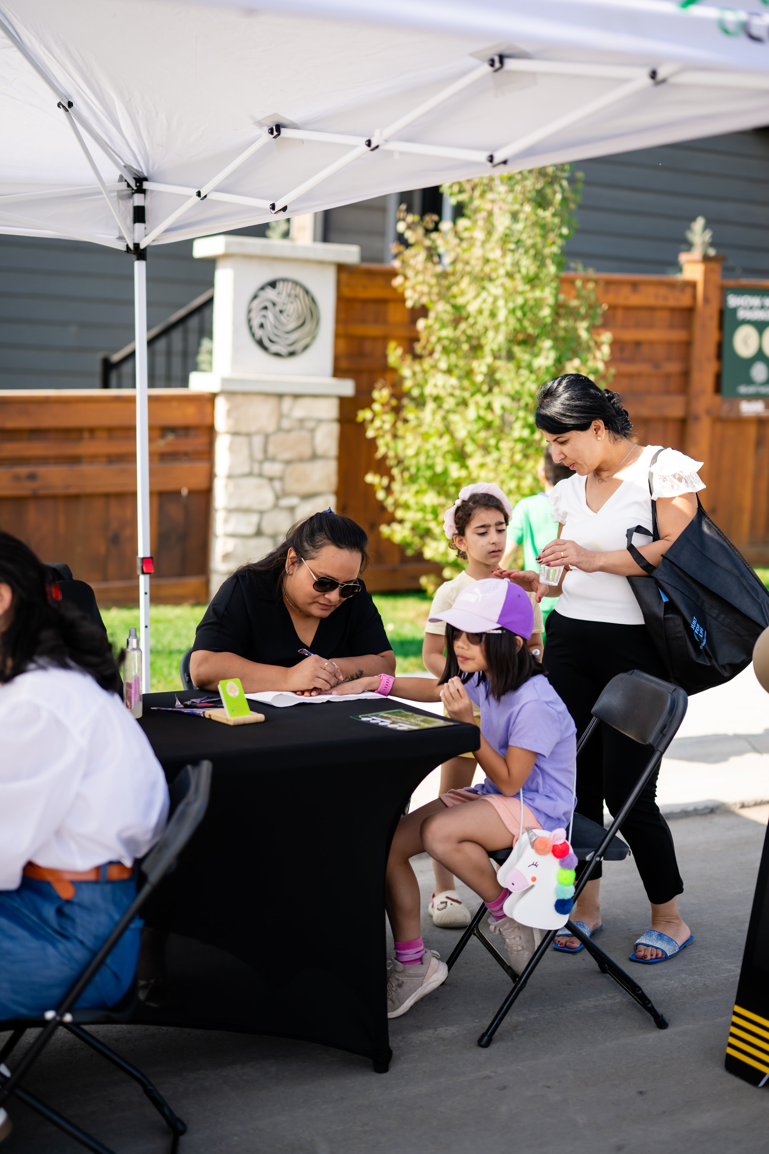 People at an outdoor table, including a woman signing an item for a young girl, under a white canopy with a black tablecloth, in a residential backyard with a wooden fence and plants.