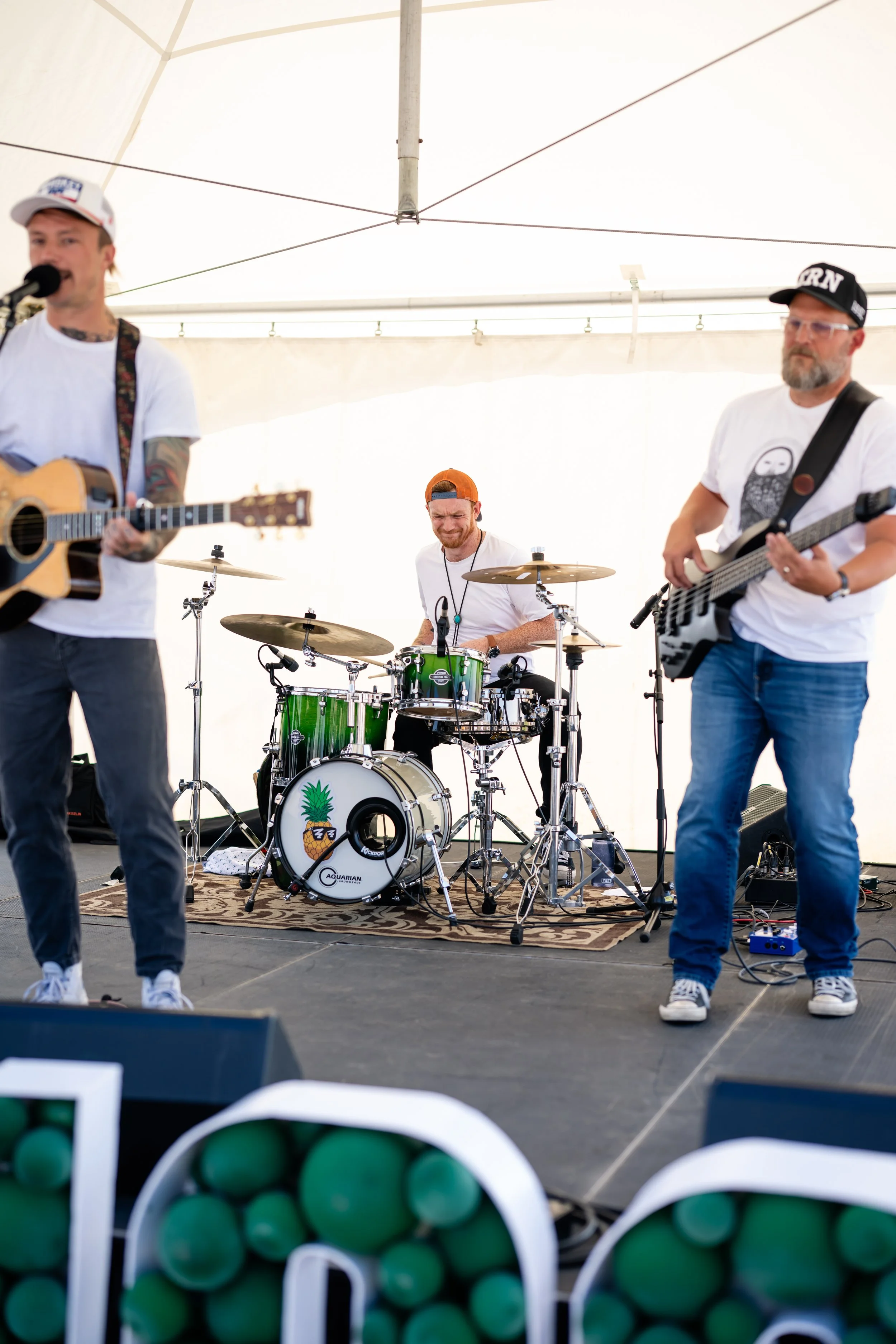 A band performing on stage in a large white tent. The band includes a guitarist, a drummer, and a bassist, all dressed casually. There are some green objects in the foreground, possibly balloons or decorations.