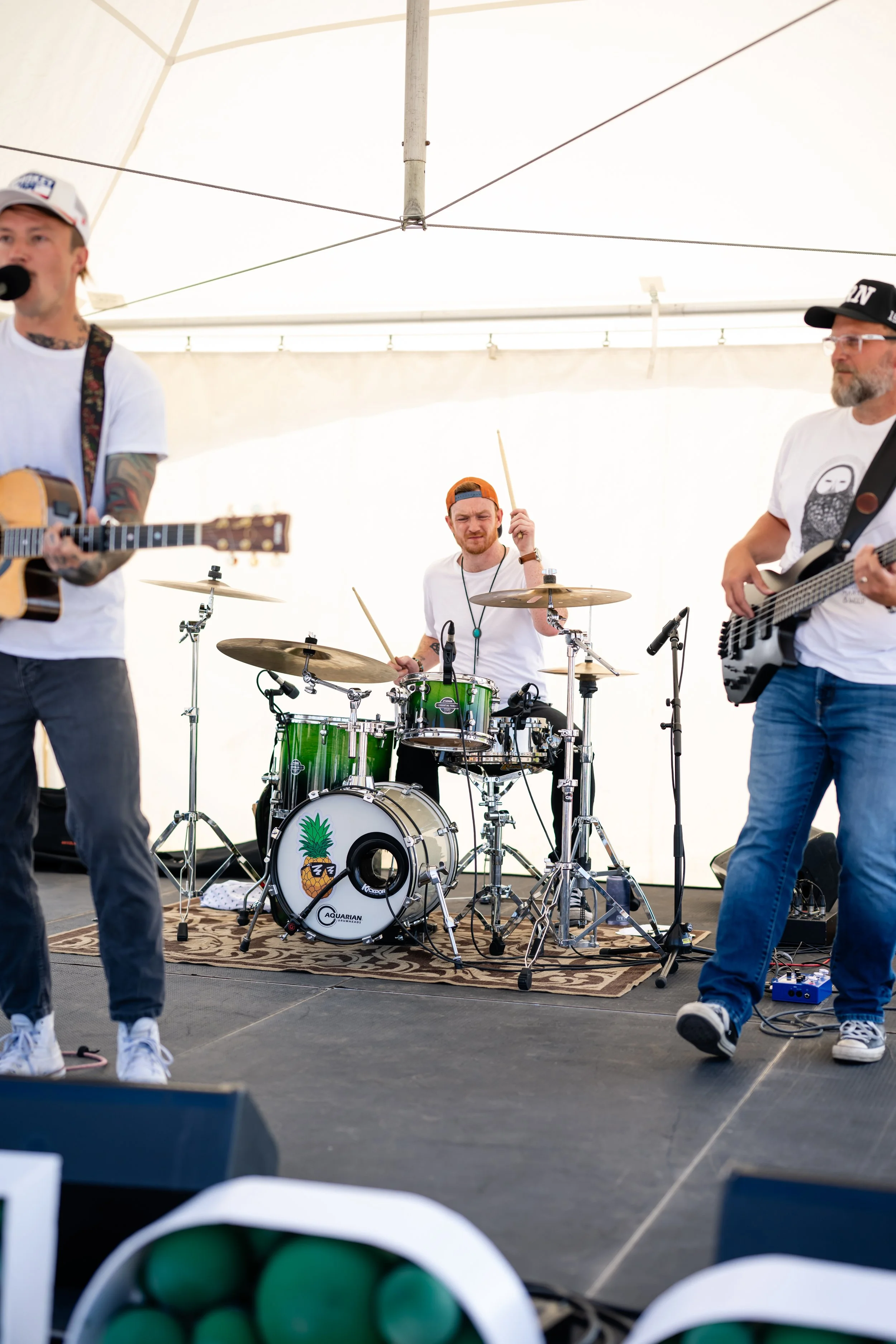 A band performing under a tent with a guitarist, drummer, and bassist playing.