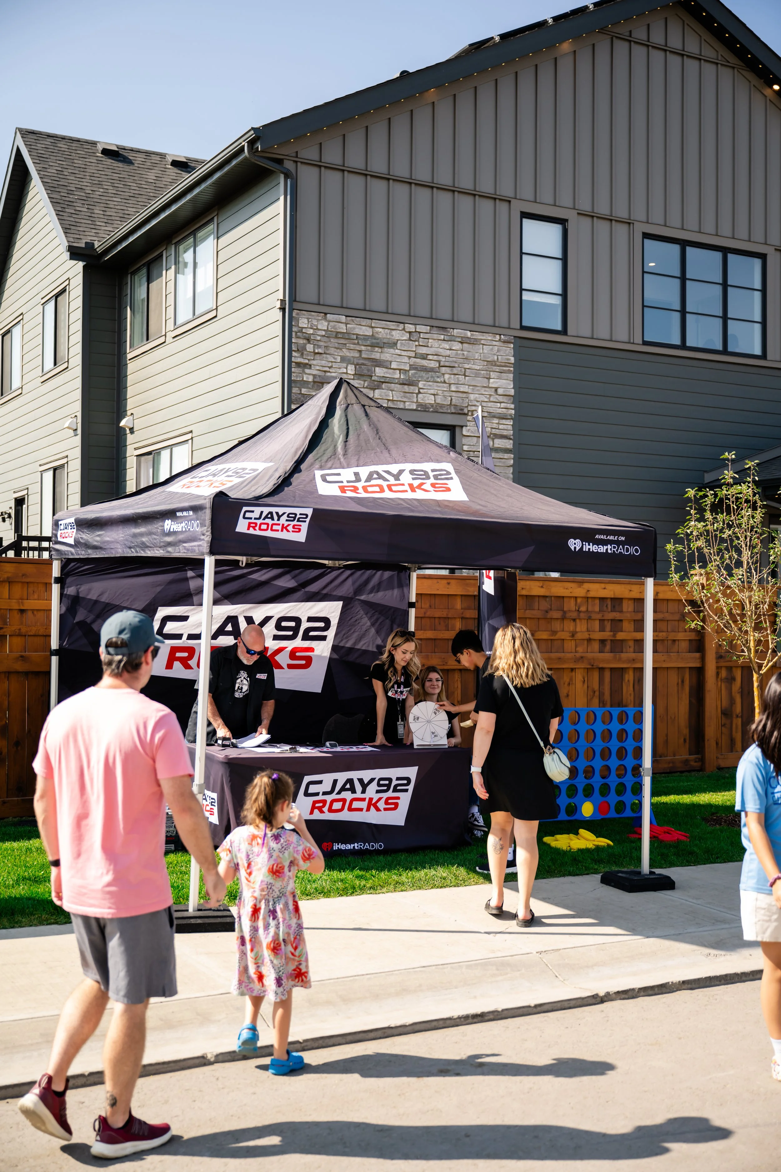 A group of people gathered around a tent with the logo of CJAY 92 ROCKS and iHeartRadio, set up outdoors in a residential neighborhood during daytime, with houses and a wooden fence in the background.