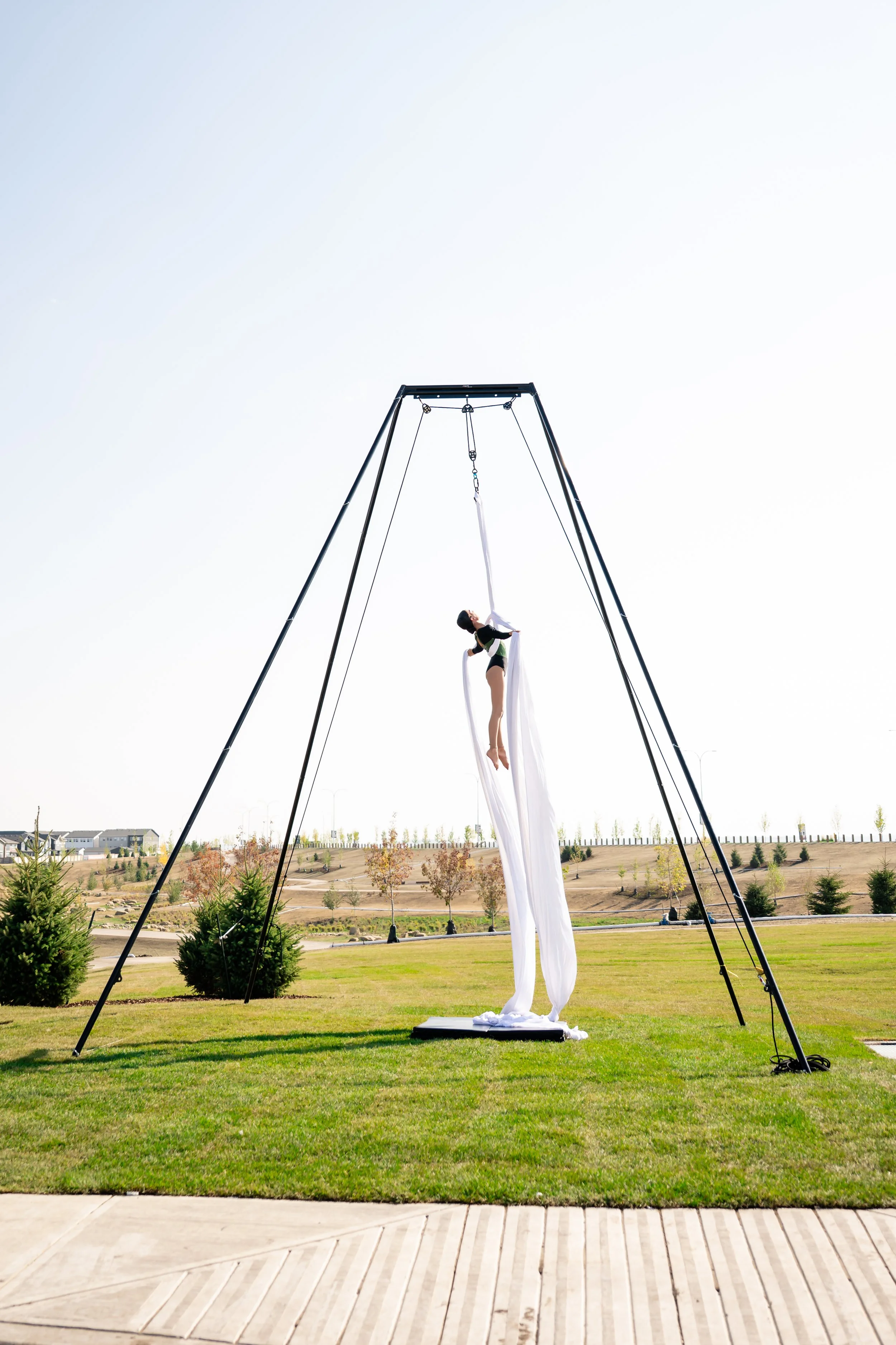 An outdoor aerial silk performance setup with a performer hanging from white silk fabric in a park area with green grass, trees, and hills in the background, and a clear sky.