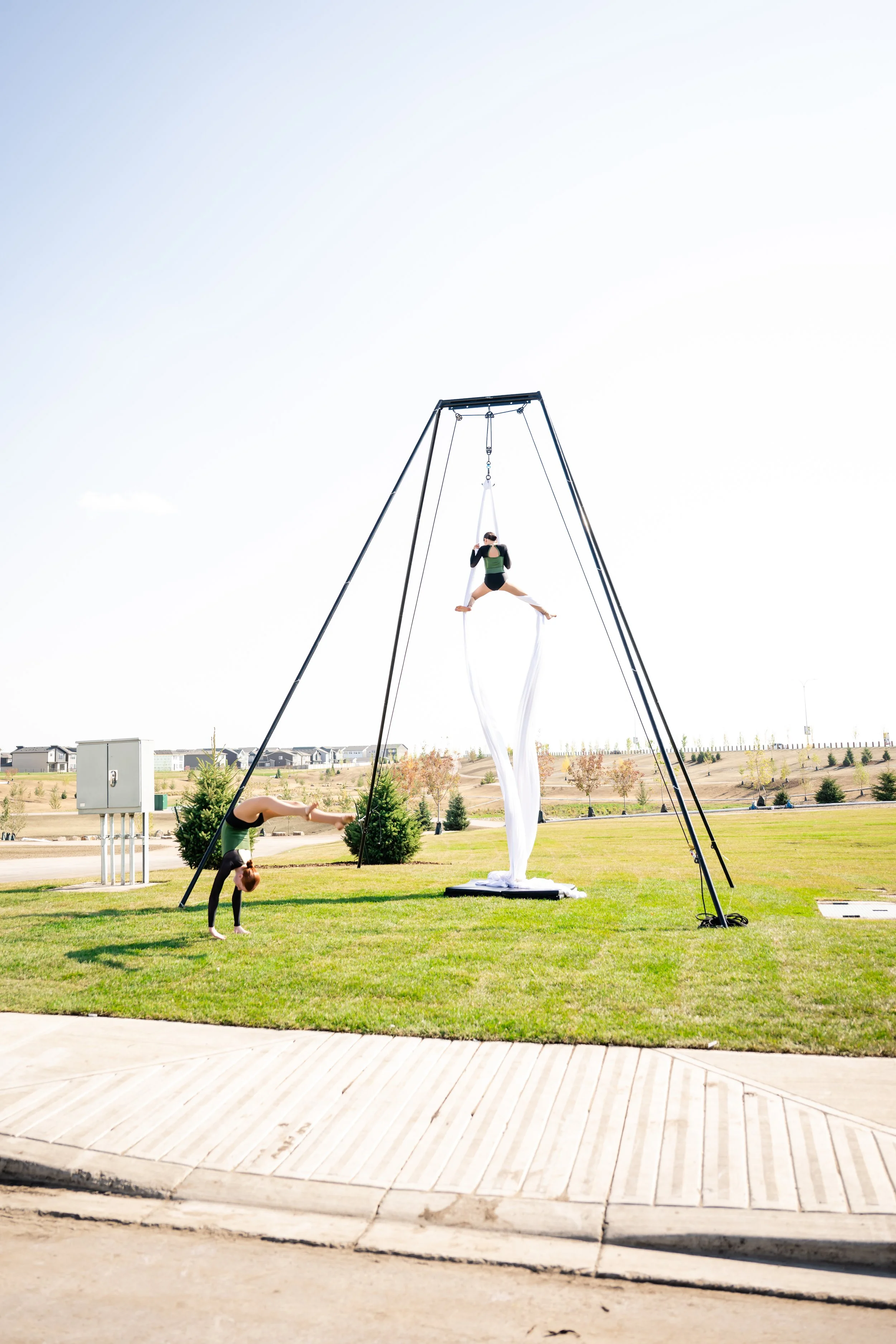 Two acrobats practicing silk performance outdoors on a grassy area. One is hanging upside down on a silk fabric with one hand grabbing their ankle, while the other is suspended in a split on the silk. A large metal frame supports the silk, and a thir