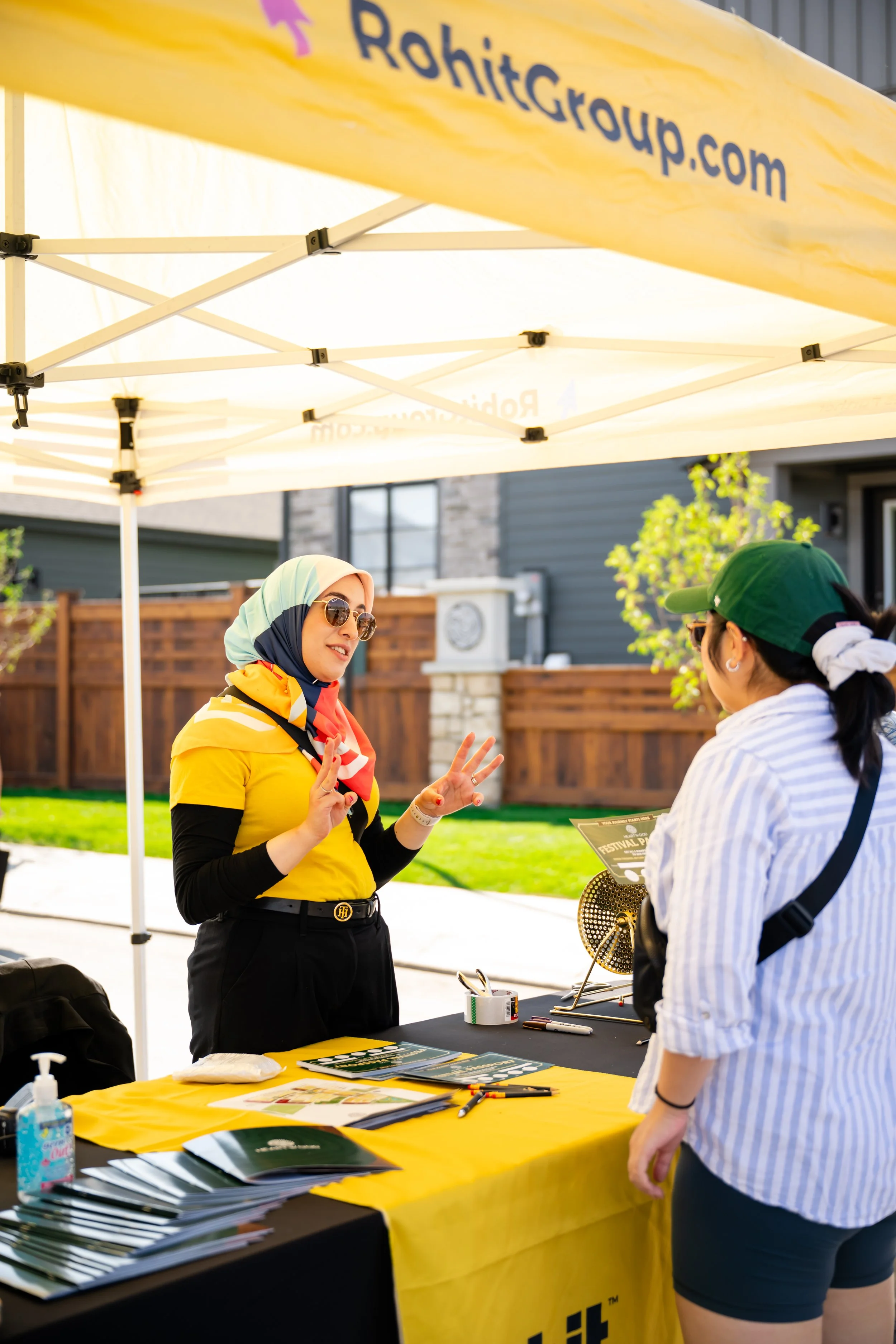 A woman wearing sunglasses and a yellow shirt with a colorful scarf is talking to another woman at an outdoor table under a yellow canopy with the logo and website of Rohit Group. The table has brochures, a bundle of pens, and a flyer, with a bottle 