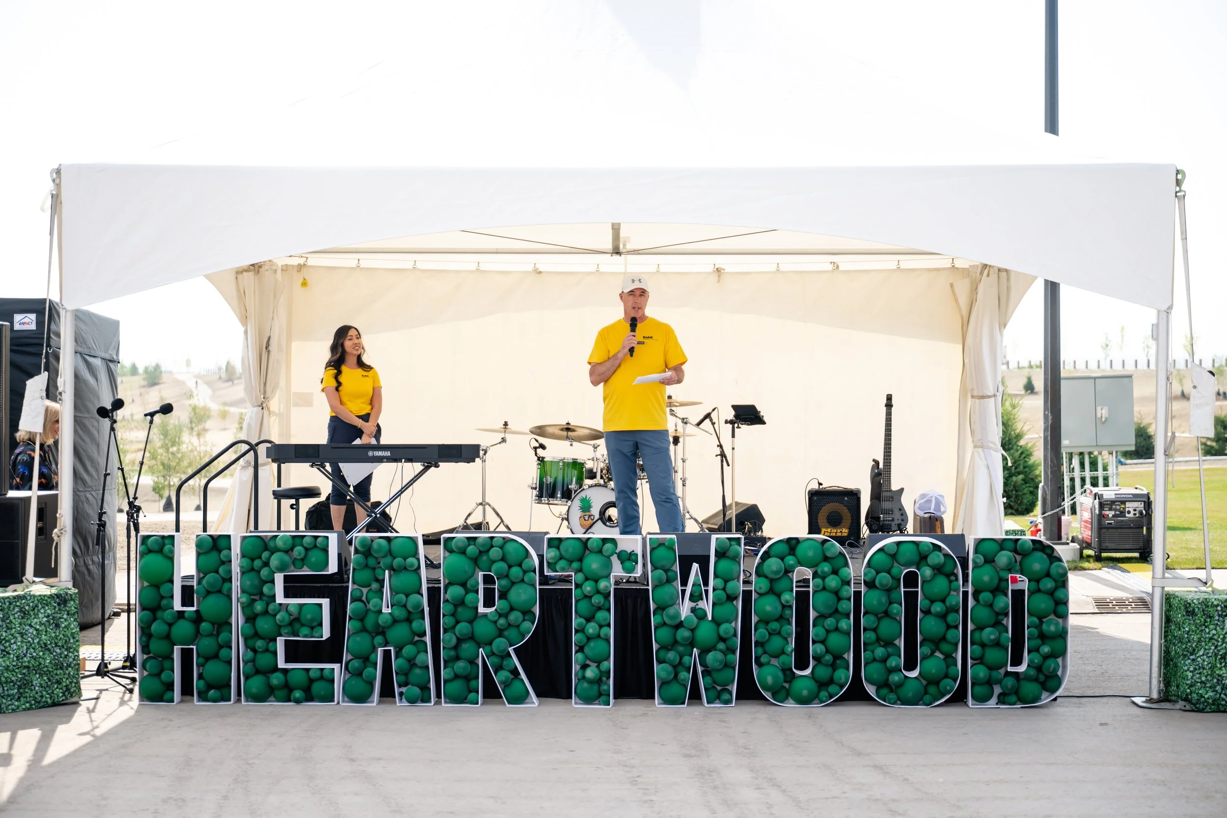 A man in a yellow shirt and white cap speaking into a microphone on a stage with musical instruments, including a keyboard and drum set, in an outdoor covered area. A young woman in a yellow shirt stands behind him. Large illuminated letters spelling