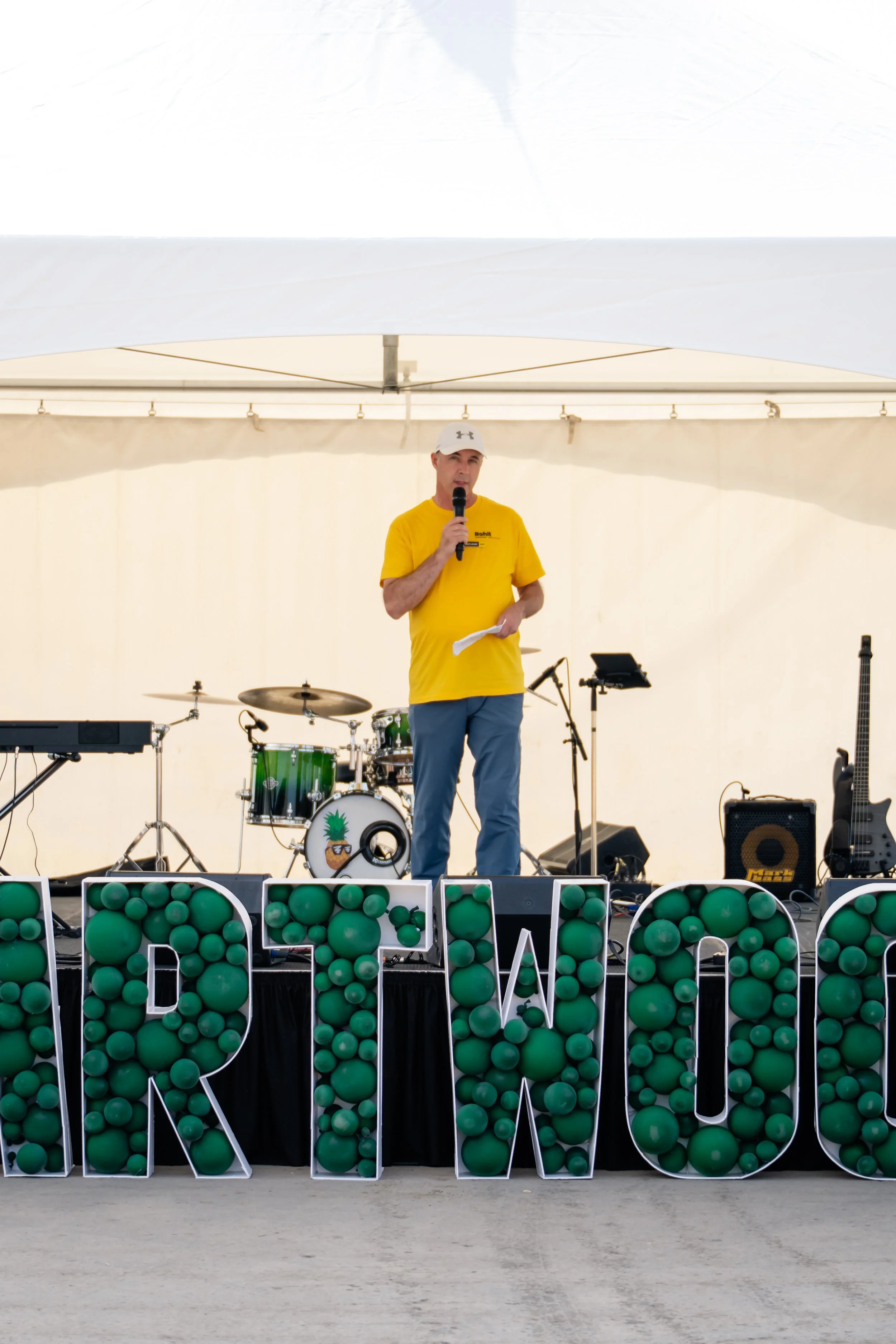 Man in yellow shirt and white cap holding microphone and paper, standing in front of musical instruments and large decorative letters on stage at outdoor event.