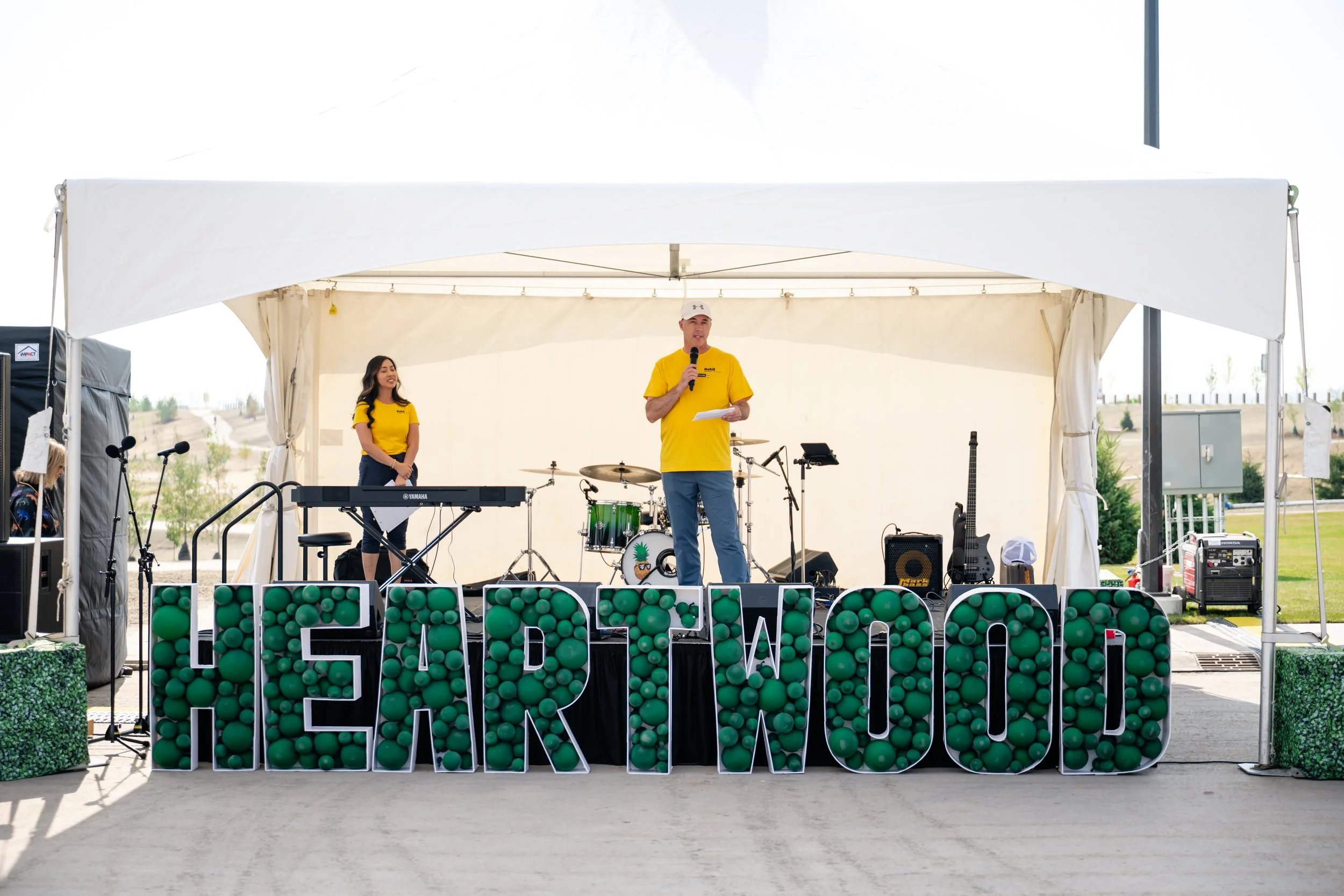 A man in a yellow shirt and white cap speaks into a microphone on a stage decorated with green and white letters and green balls, with a woman in a yellow shirt standing nearby. The stage is set up outdoors under a white tent with musical instruments