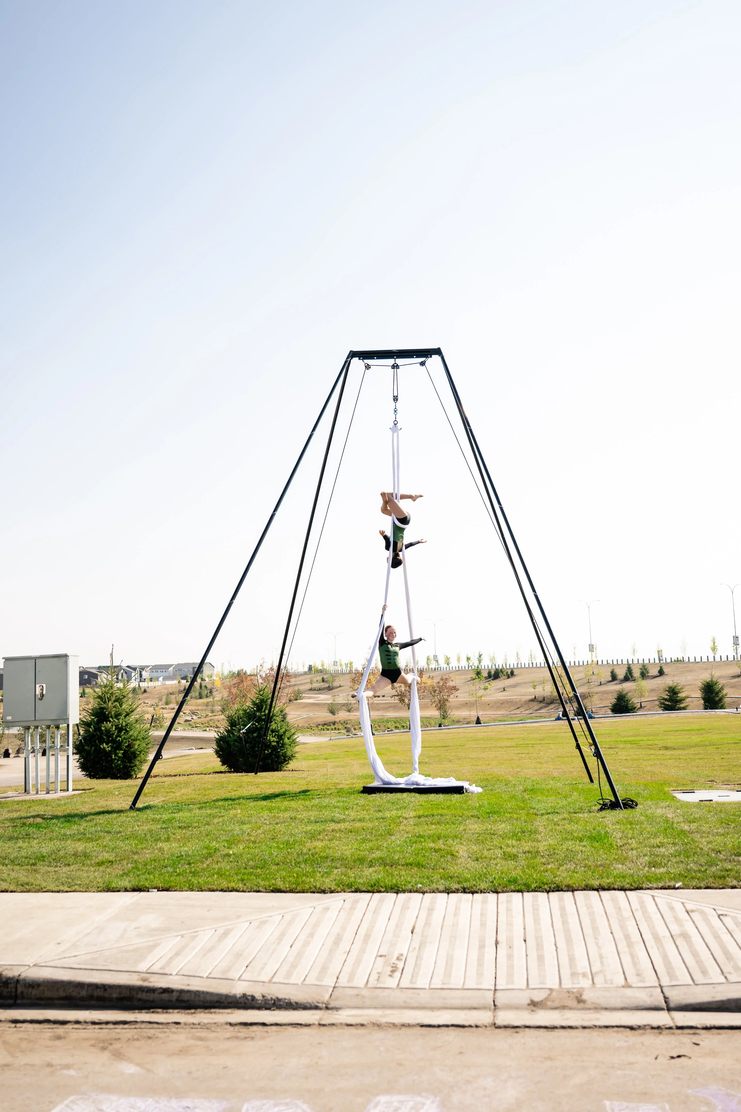 Two aerialists performing on white silks in an outdoor park, with a large trapeze structure behind them and houses in the background.