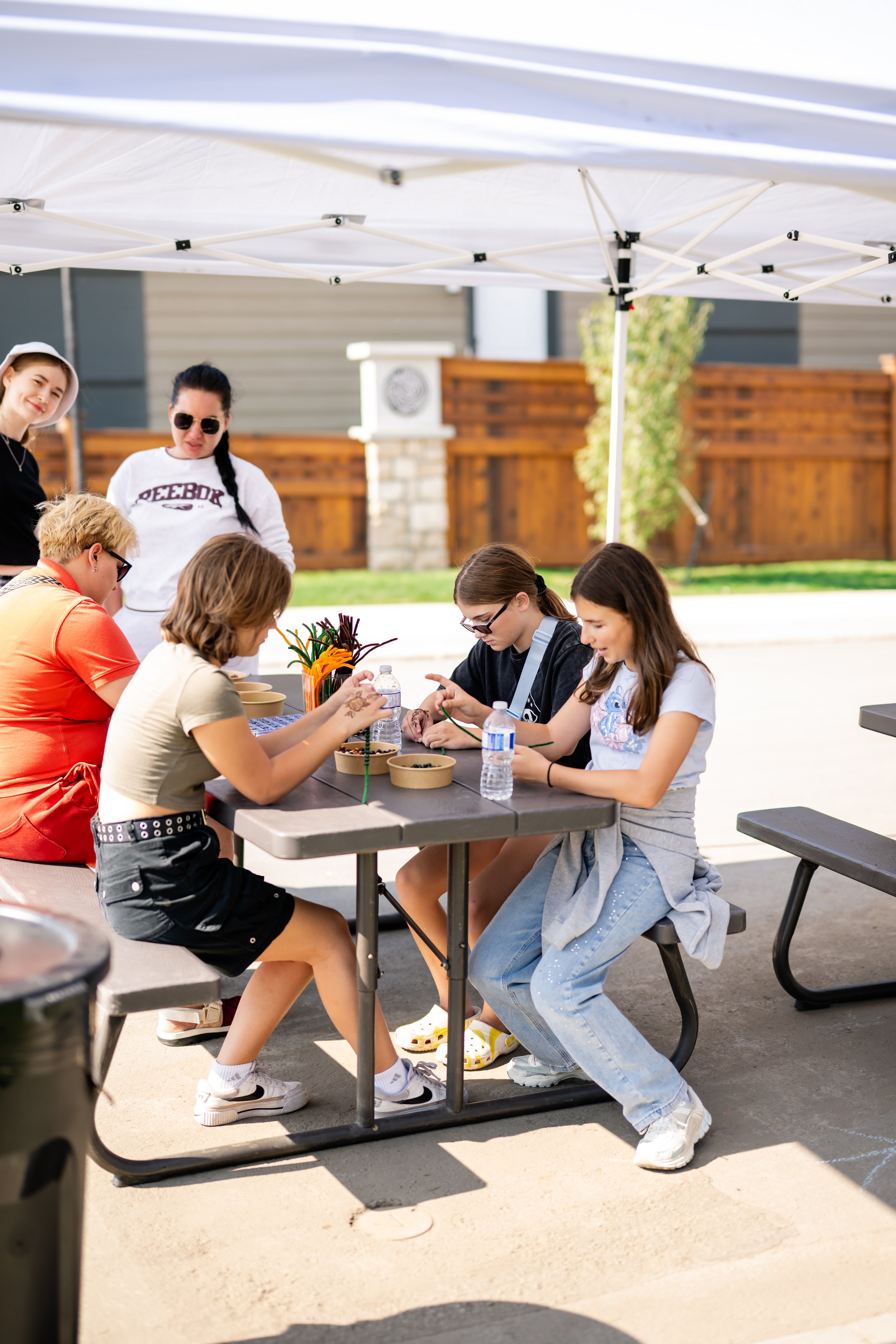 Children and adults decorating flower pots outdoors under a white canopy, with a wooden fence and green plants in the background.