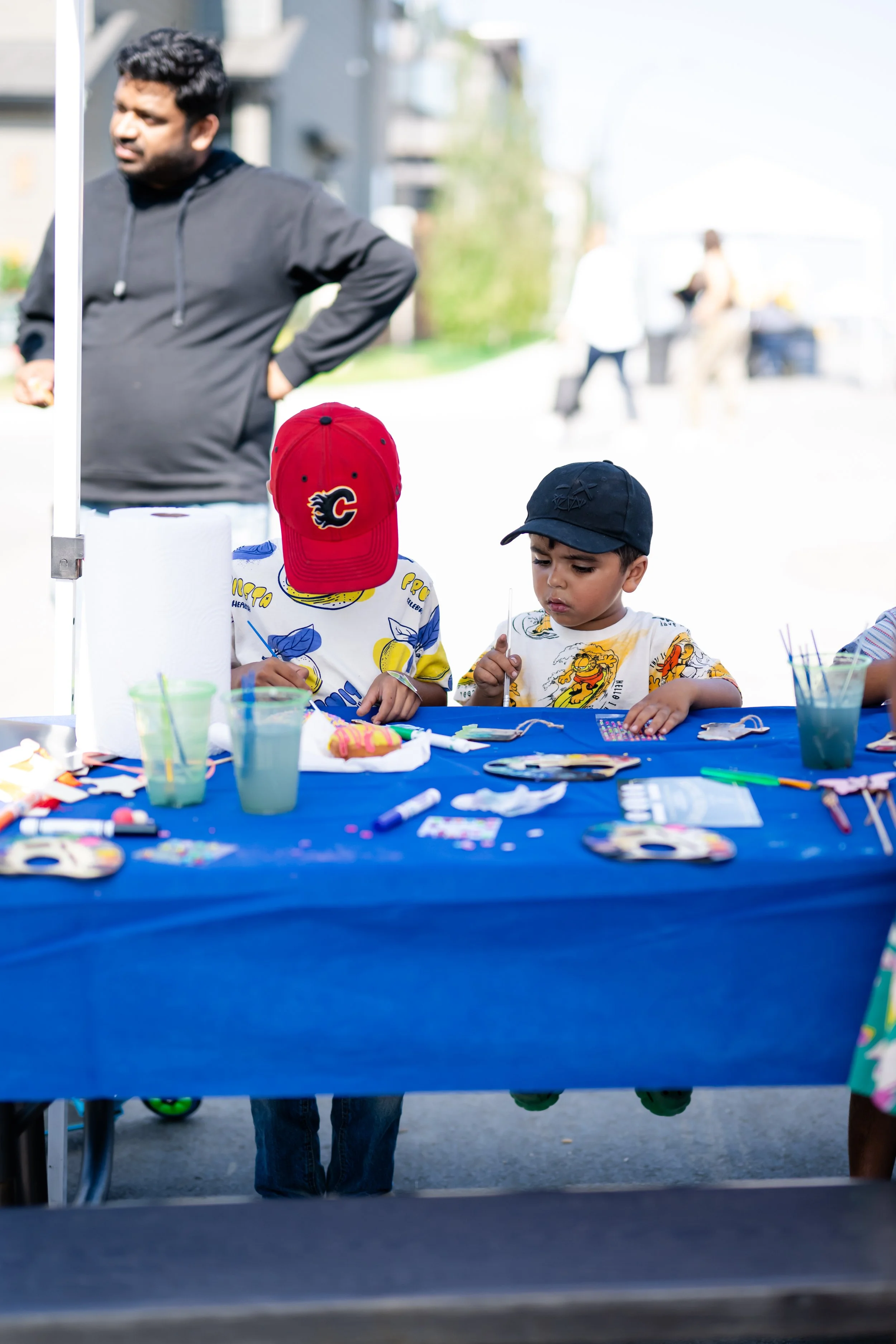 Two children at a table participating in outdoor arts and crafts activities. One child wears a red cap with a Calgary Flames logo, and the other wears a navy cap with a design. The table is covered with various art supplies.
