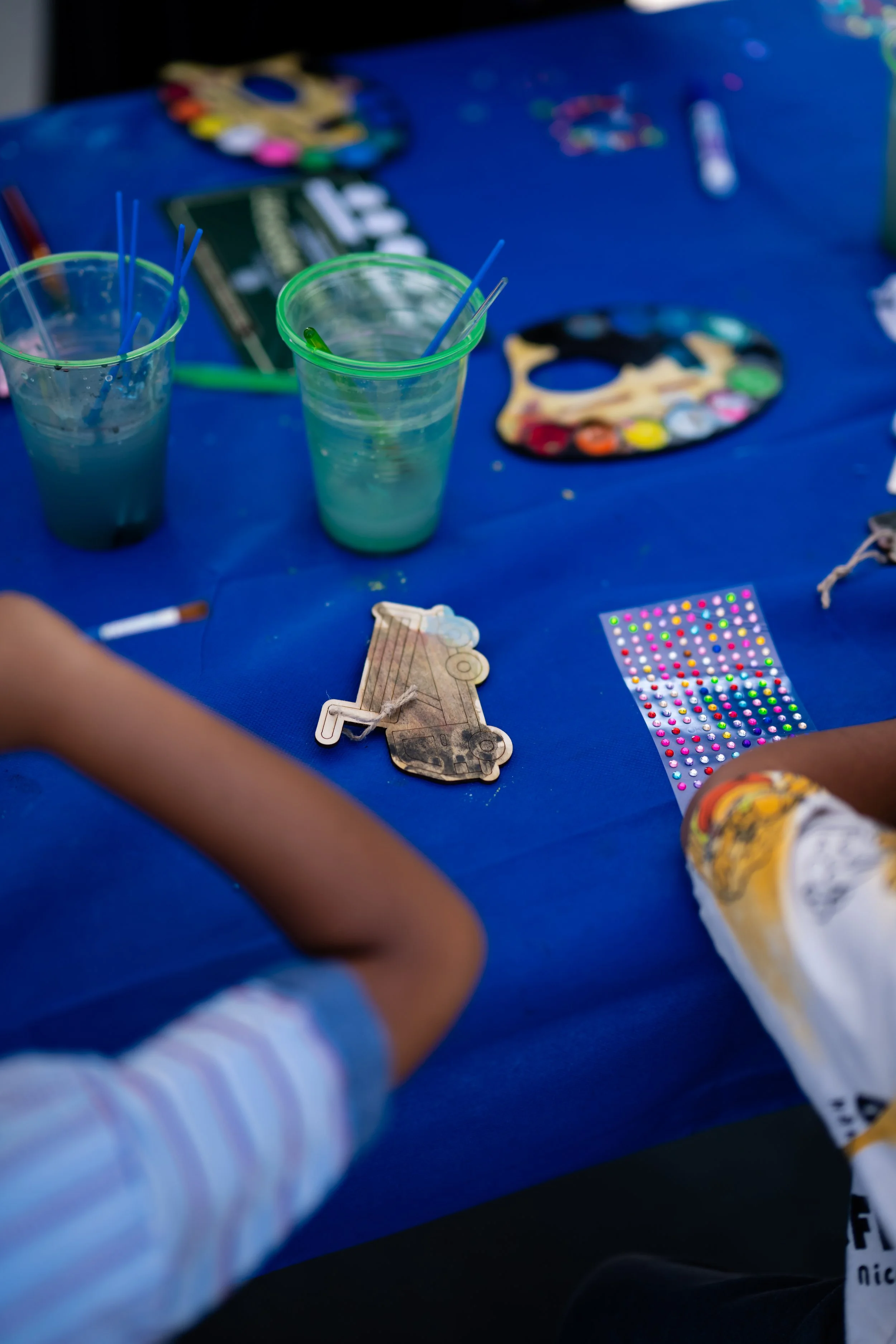 Children's craft or art activity setup with two cups of water, paintbrushes, a paint palette, and decorative stickers on a blue tablecloth.