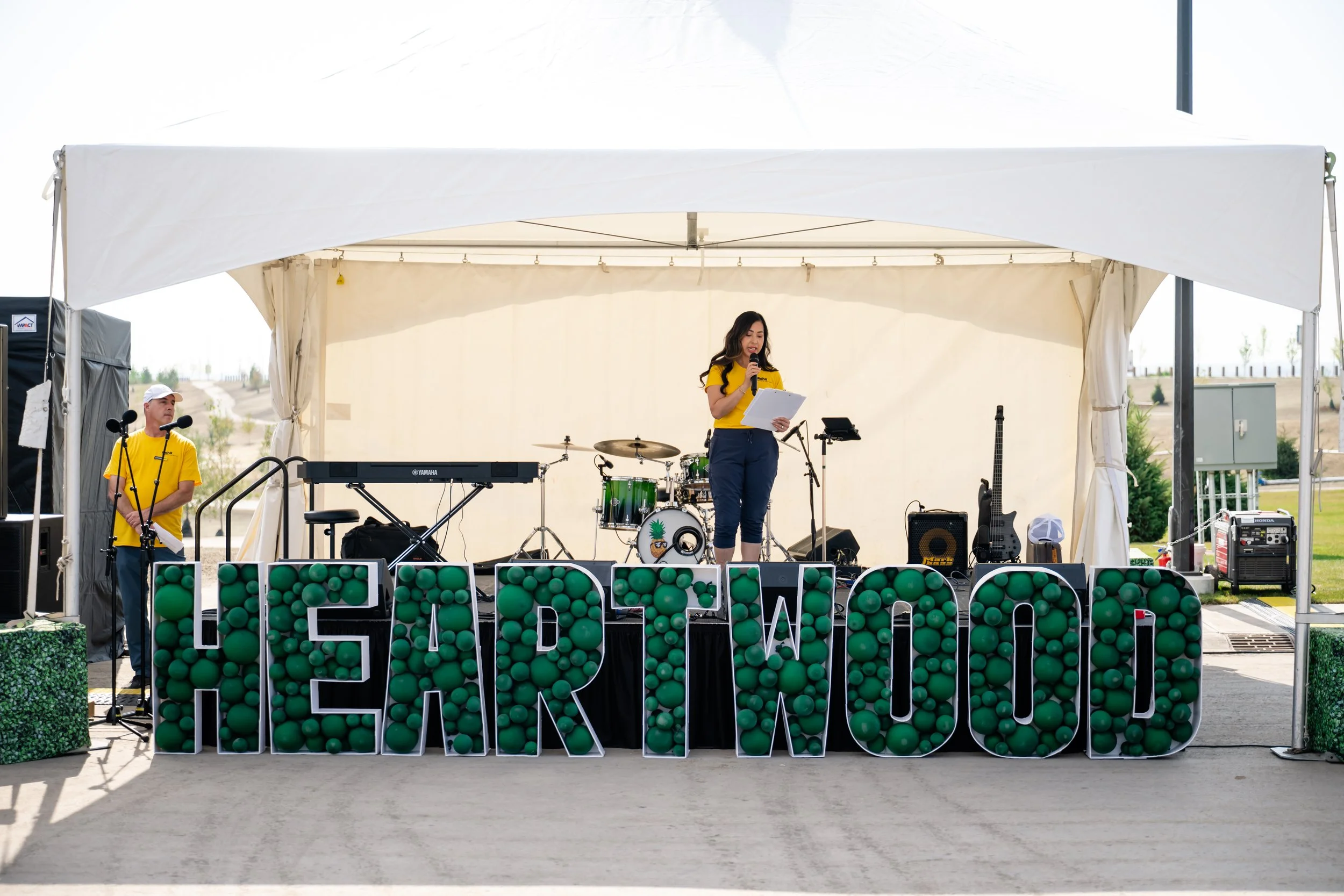 A woman with dark hair in a yellow shirt reads from a paper on a stage with musical instruments and green decorations, outside under a white tent.