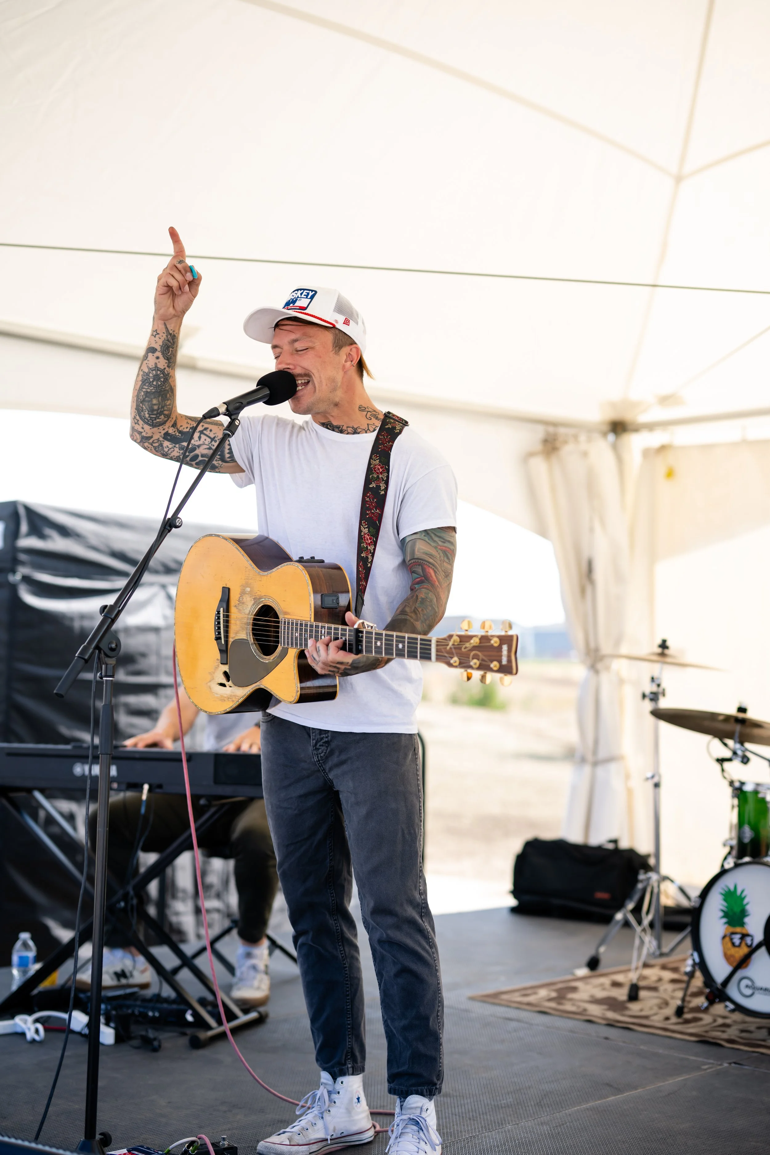 A man with tattoos singing and playing guitar under a large white tent, with musical equipment and a keyboard in the background.