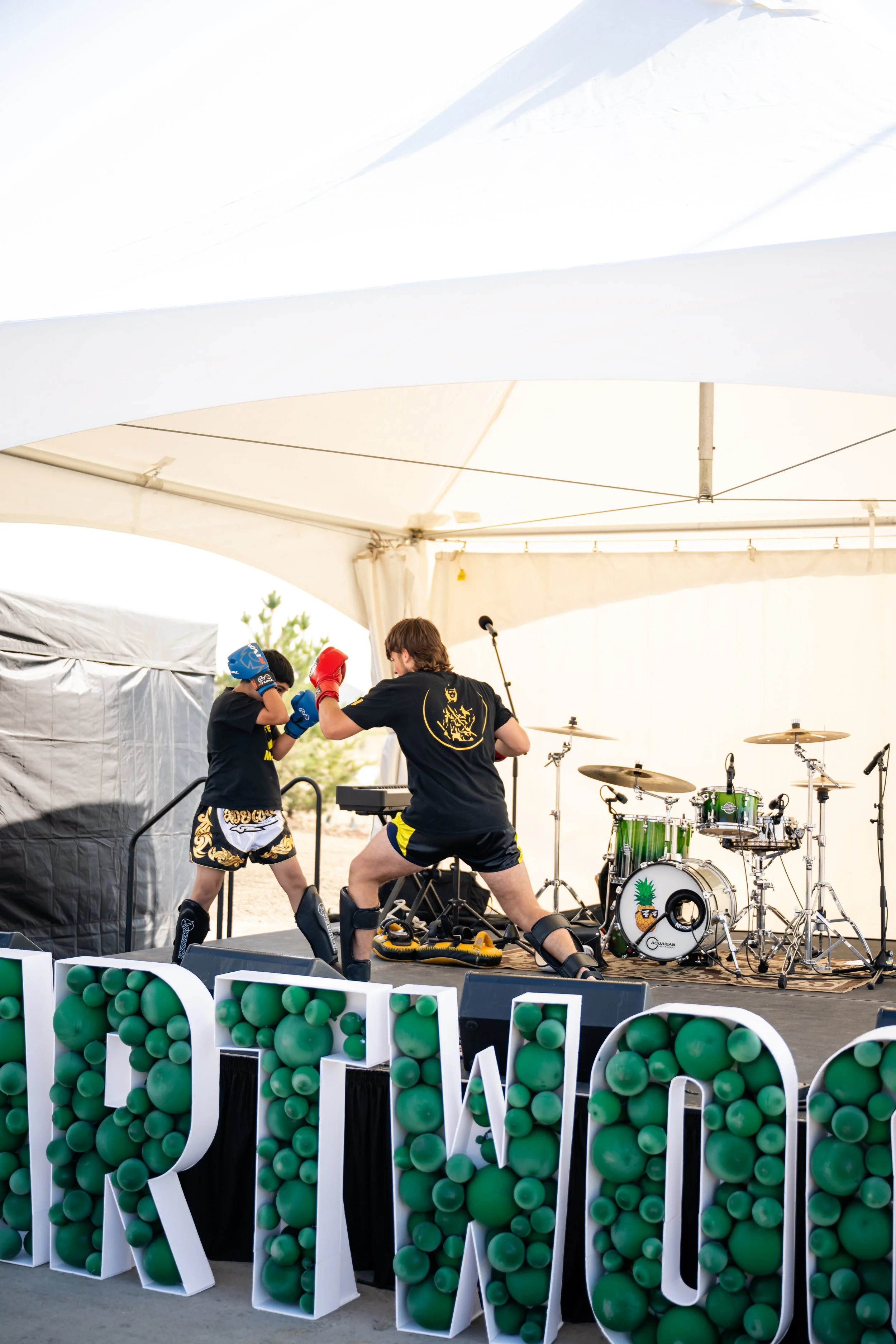 A person sparring with a young boy in a boxing glove under a white tent. There are drums and musical equipment in the background and large letters with green balls in the foreground.