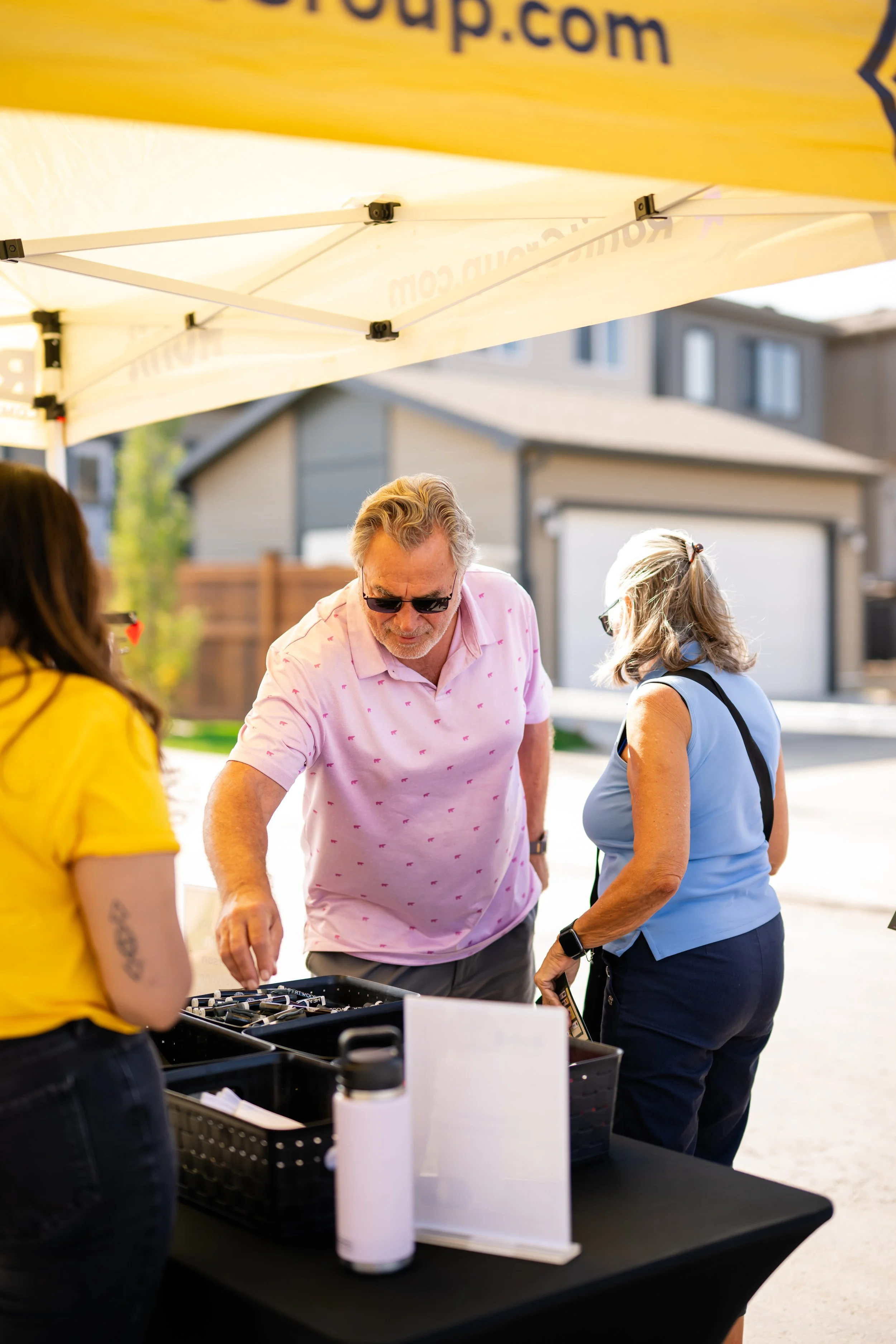 People gathered around a table under a yellow canopy at an outdoor event, with houses and trees in the background.